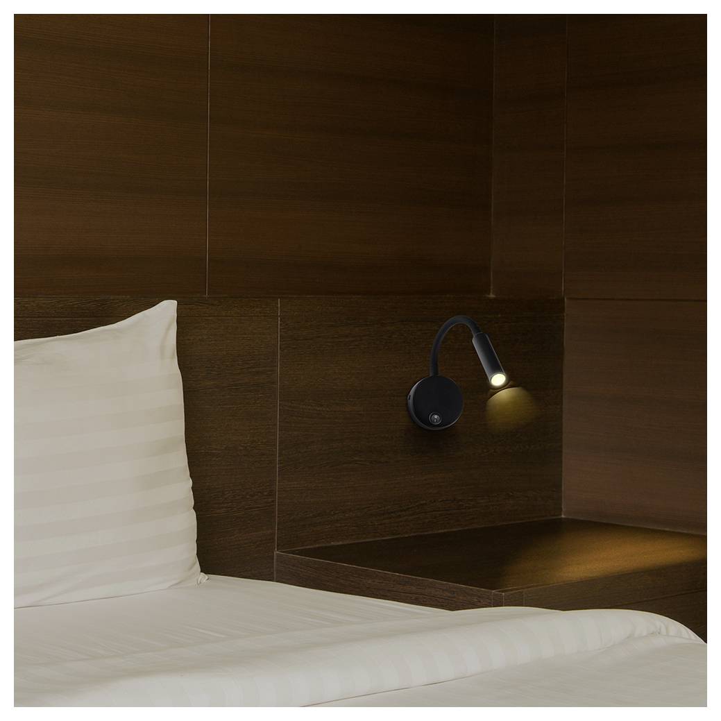Ein ordentlich gemachtes weißes Bett neben einem dunkelbraunen Holzkopfteil mit einer klein leuchtenden Leselampe, was ein gemütliches hotelartiges Schlafzimmer andeutet.