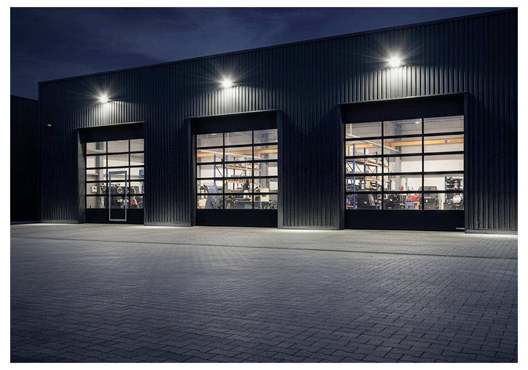 Außenseite eines modernen Industrie-Lagerhauses bei Nacht, mit drei großen beleuchteten Garagentoren, die Ausrüstung im Inneren zeigen.