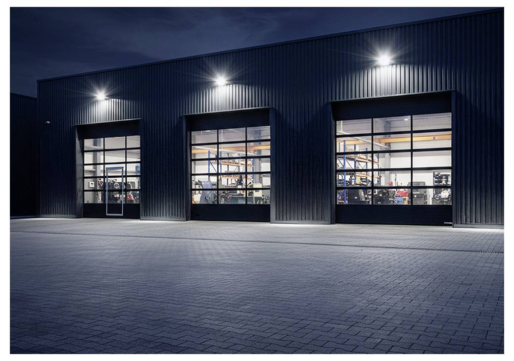 Nächtliche Außenaufnahme eines Industrielagers mit drei beleuchteten Glasschiebetoren, die Ausrüstung im Inneren zeigen, und einem leeren gepflasterten Vorplatz.