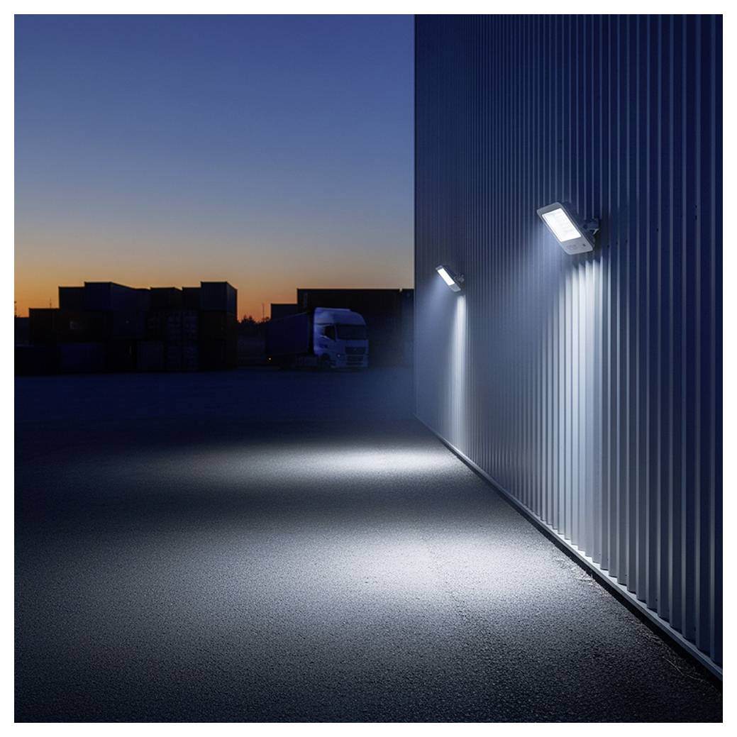 Außenseite eines Lagers in der Abenddämmerung, wobei wandmontierte Leuchten die wellblechverkleidete Wand und das Pflaster beleuchten, und ein Lastwagen im Hintergrund als Silhouette zu sehen ist.