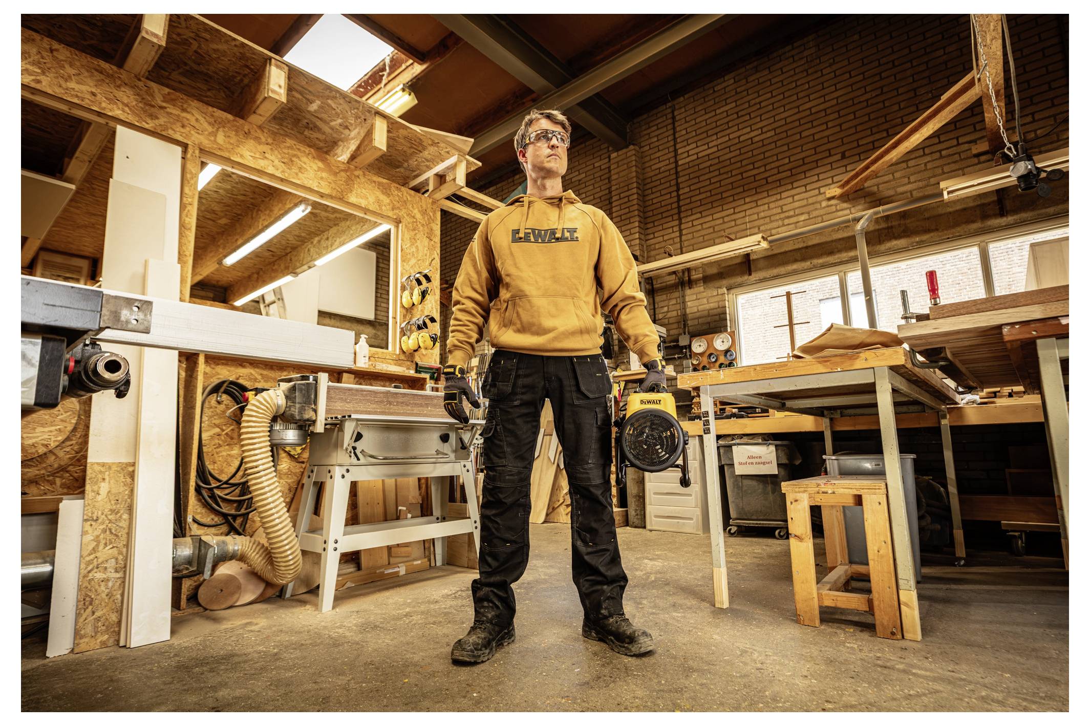 Ein Mann in einer Werkstatt steht und hält ein Elektrowerkzeug, umgeben von Holzbearbeitungsgeräten und Werkbänken in einem Geschäft im Industriestil.