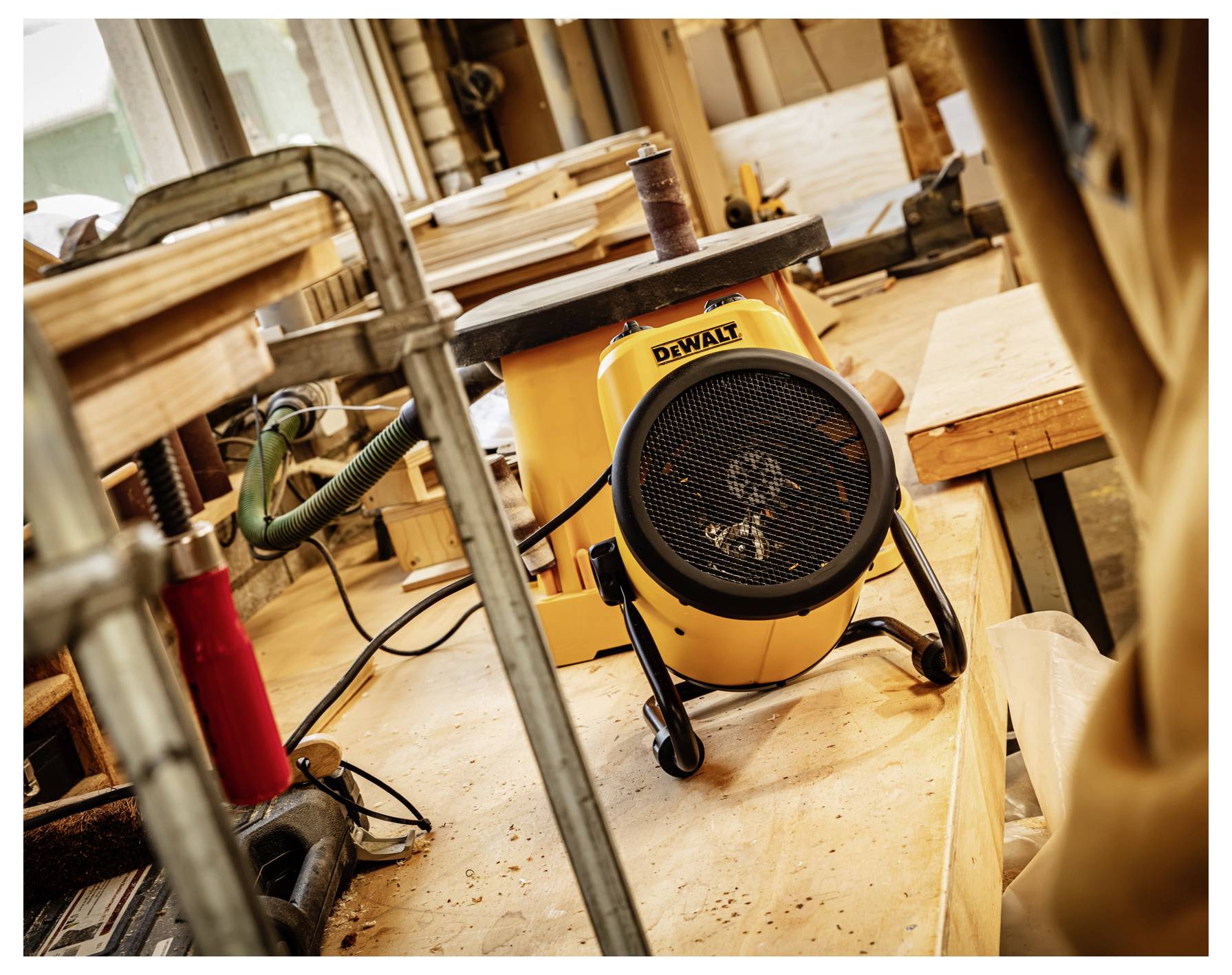 Tragbarer gelber DeWalt-Elektroheizer auf einem Werkstattboden zwischen Holz und Werkzeugen, warme Luft in einem Bau- oder Zimmermannsbereich blasend.