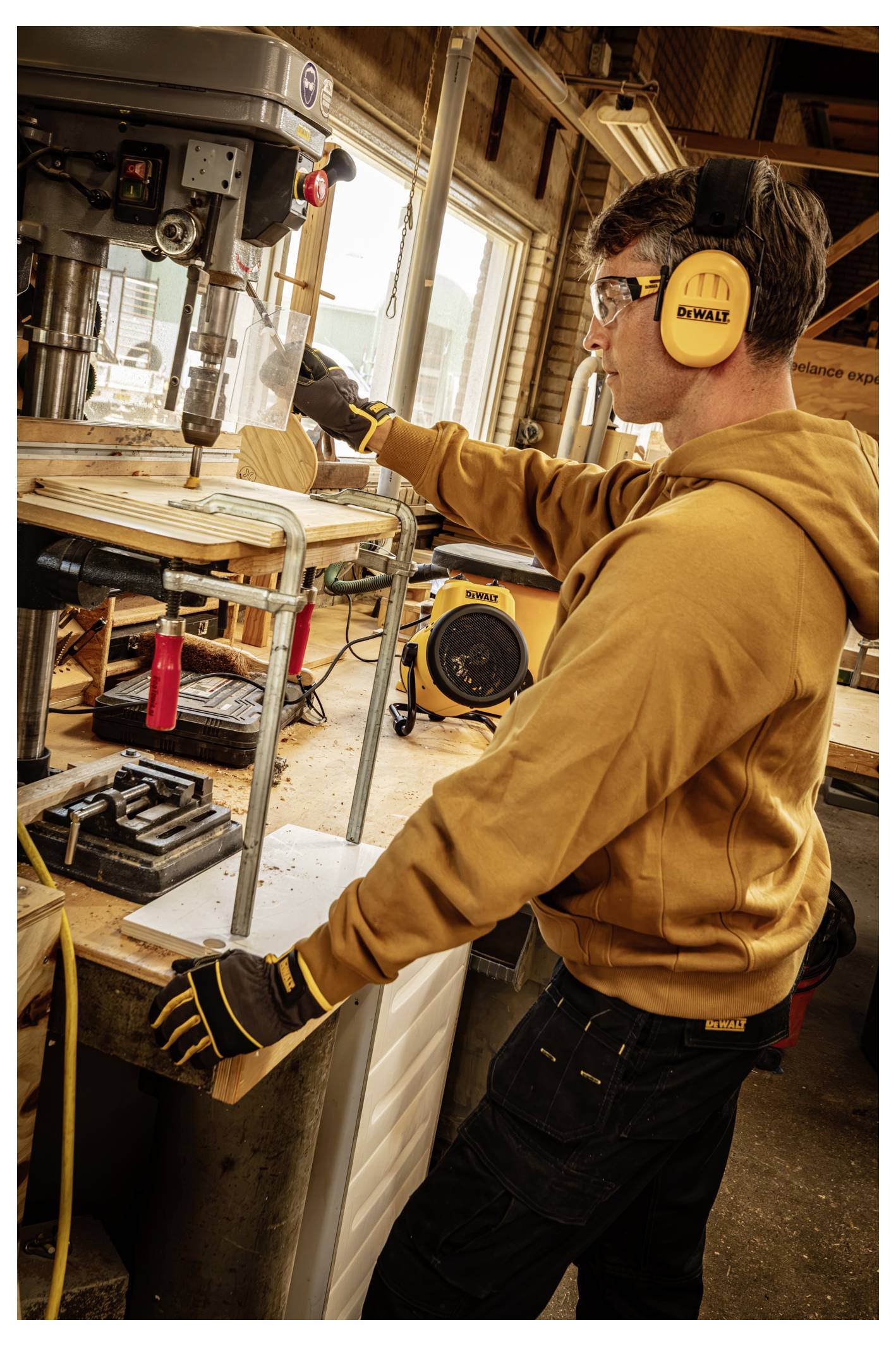 Mann bedient eine Bohrmaschine in einer Werkstatt, trägt eine Schutzbrille, gelben Gehörschutz, Handschuhe und einen Kapuzenpullover, konzentriert sich auf das Bohren von Holz.