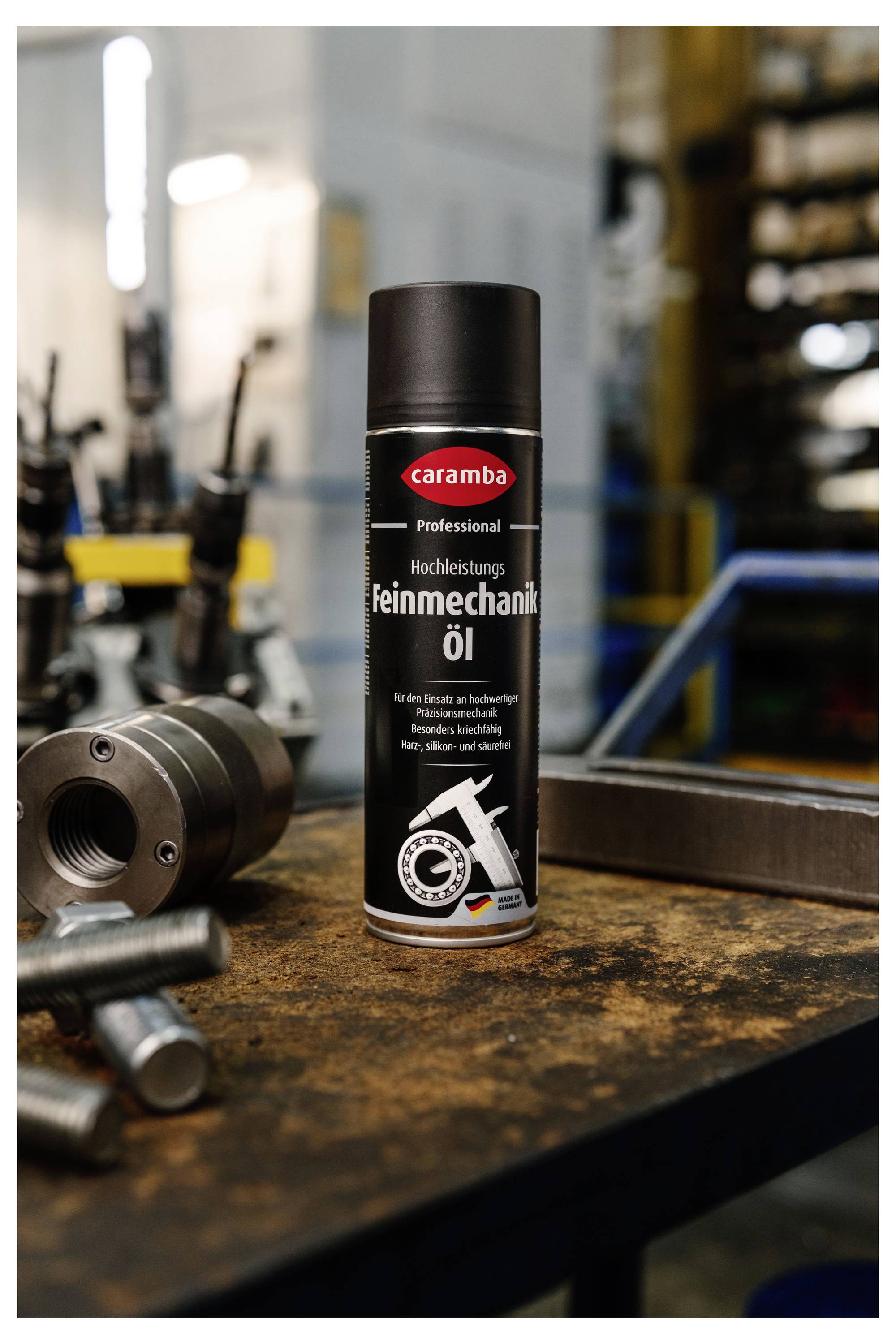 Eine Dose Caramba Professional Hochleistungs-Präzisionsöl steht auf einem Werkstatttisch neben Metallteilen, mit unscharfem Industriehintergrund.