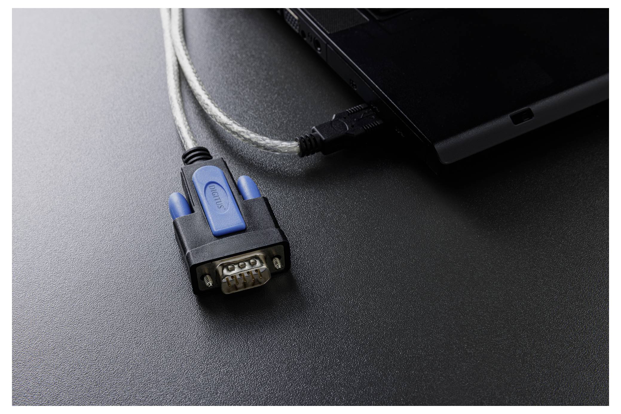 Nahaufnahme eines Laptops mit einem angeschlossenen Seriell-zu-USB-Adapter und Kabel auf einer dunklen Oberfläche.