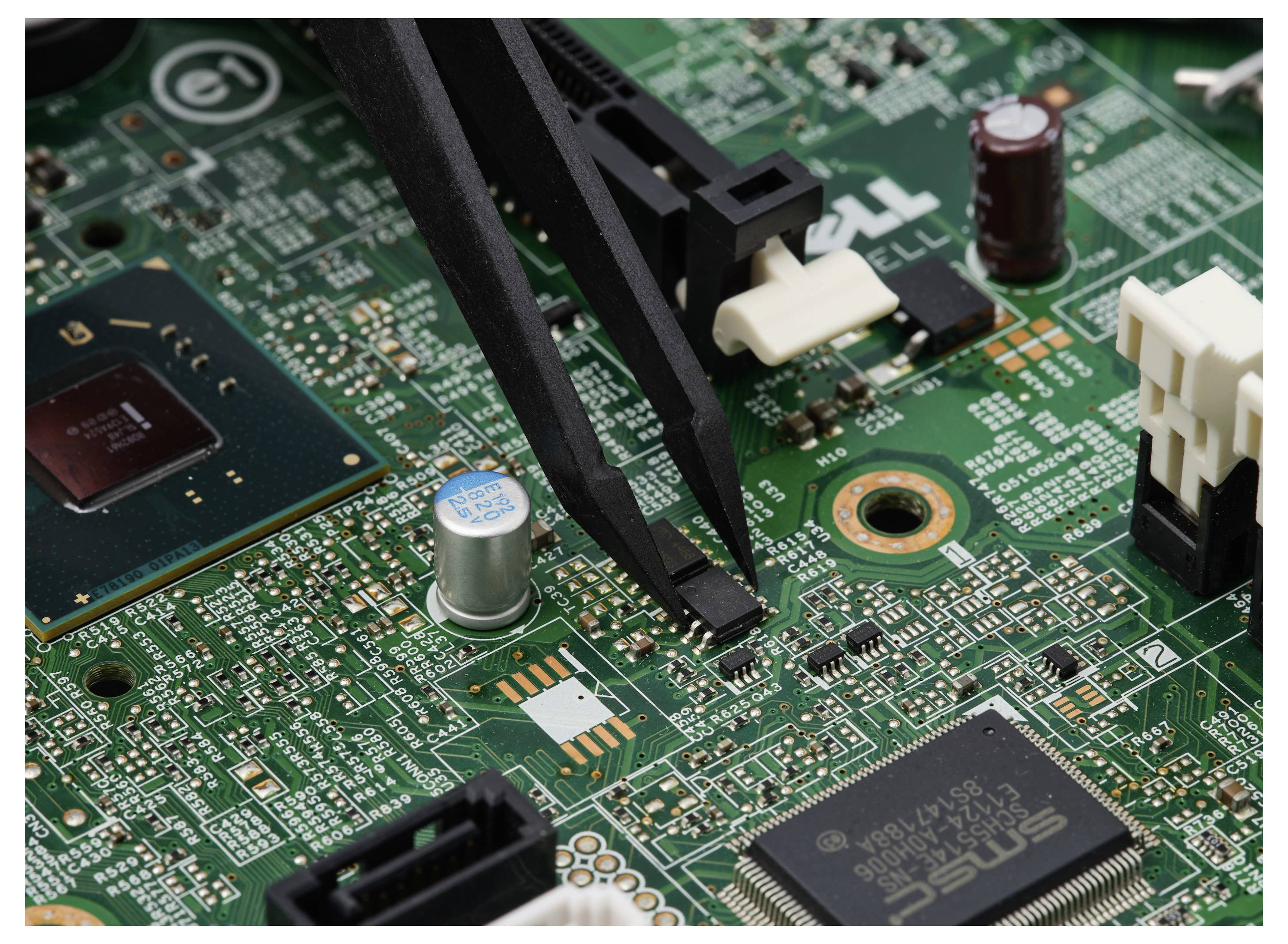 Nahaufnahme einer Computerplatine mit einem Paar schwarzer Pinzetten, die eine winzige oberflächenmontierten Komponente in der Nähe der Mitte handhaben.