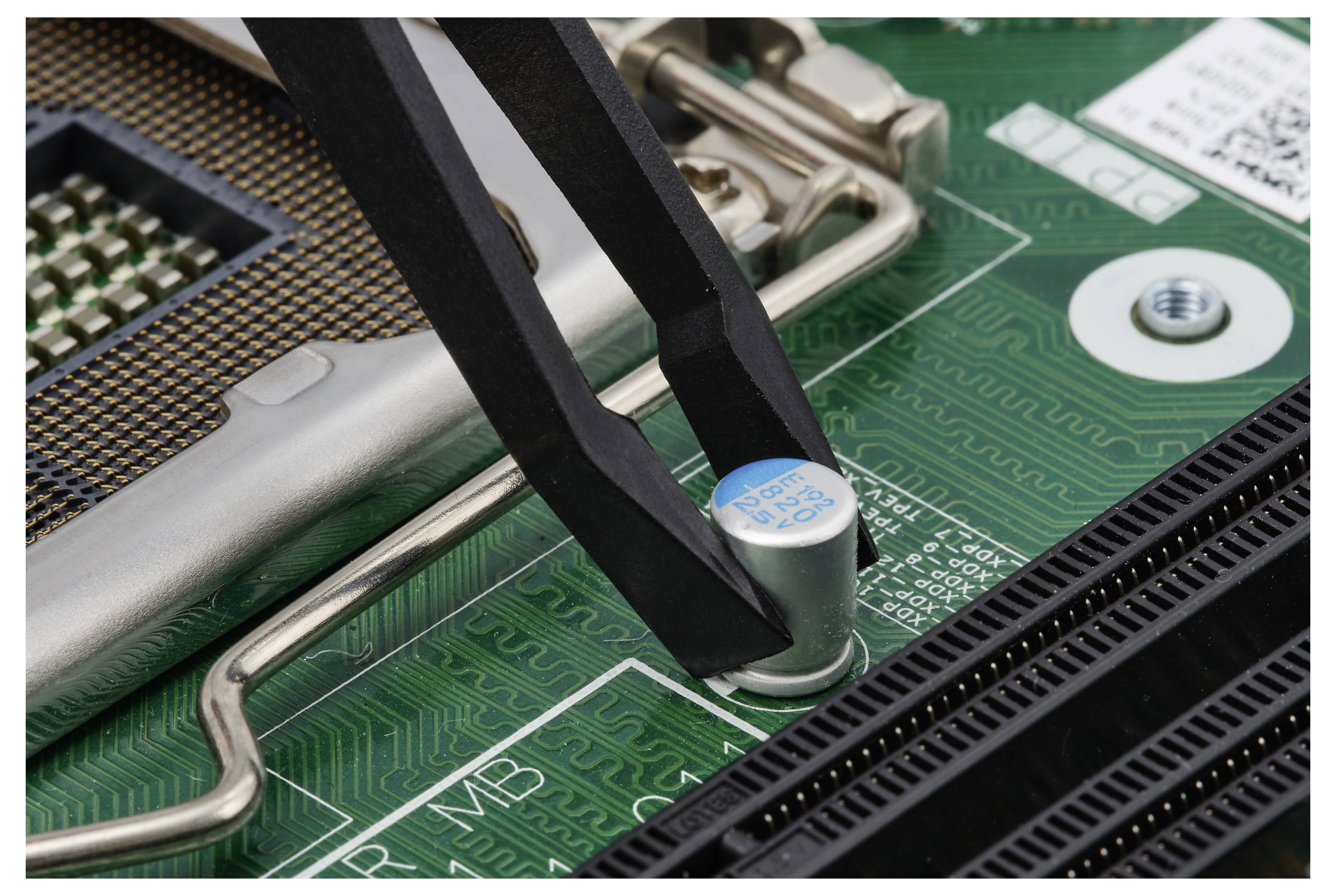 Nahaufnahme von Computerplatinen-Komponenten mit einer schwarzen Klammer, die einen Kondensator mit blauem Deckel in der Nähe des CPU-Sockels hält.