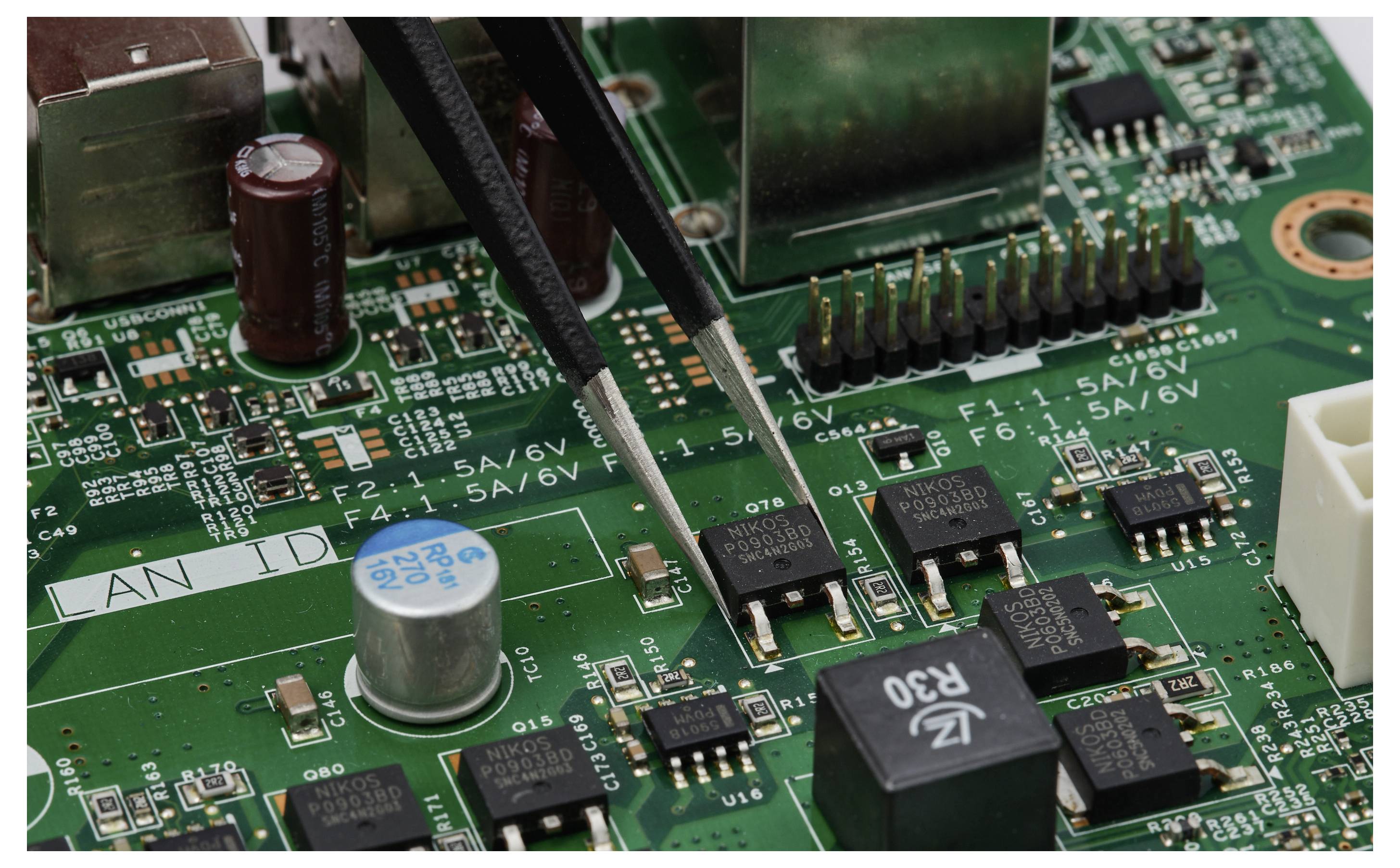 Nahaufnahme einer Leiterplatte mit Pinzette, die auf elektronische Komponenten zeigt und eine Reparatur oder Überprüfung andeutet.