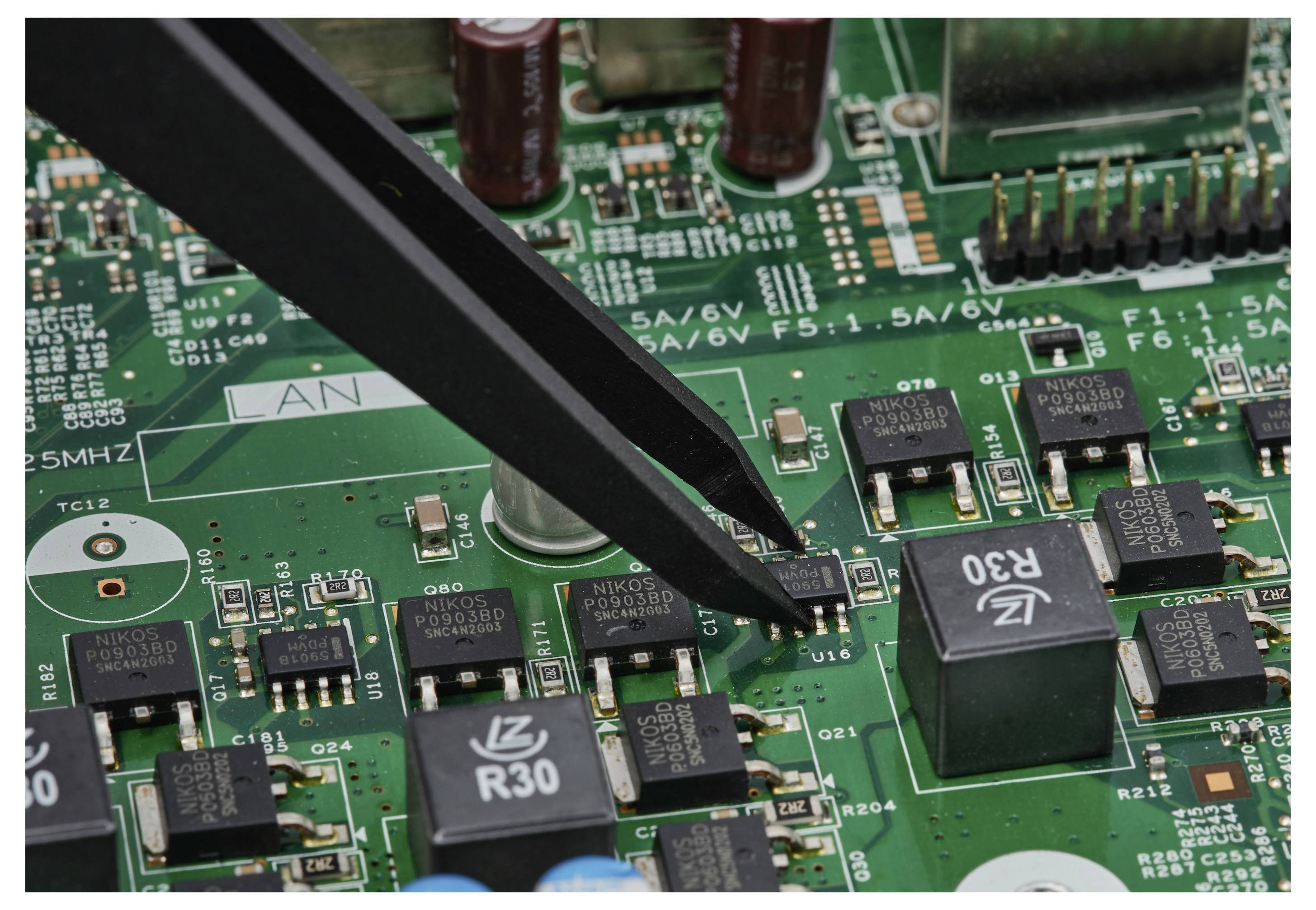 Eine Pinzette greift einen winzigen Oberflächenmontage-Chip auf einer grünen Leiterplatte während der Elektronikreparatur oder -montage.