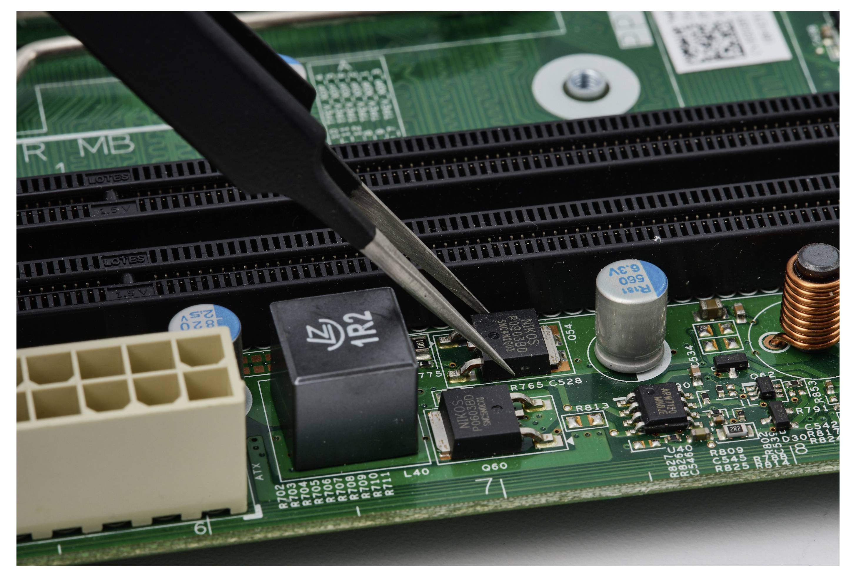 Pinzetten positionieren ein kleines elektronisches Bauteil auf einer grünen Leiterplatte neben einem schwarzen Stecker, Kondensatoren und einer Kupferspule.