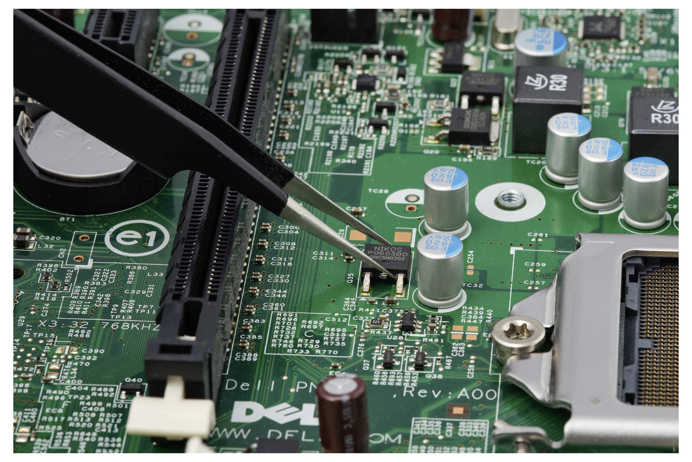 Pinzette hält einen winzigen Chip auf einer grünen Leiterplatte in einem Computer, mit sichtbaren Kondensatoren und einem CPU-Sockel.