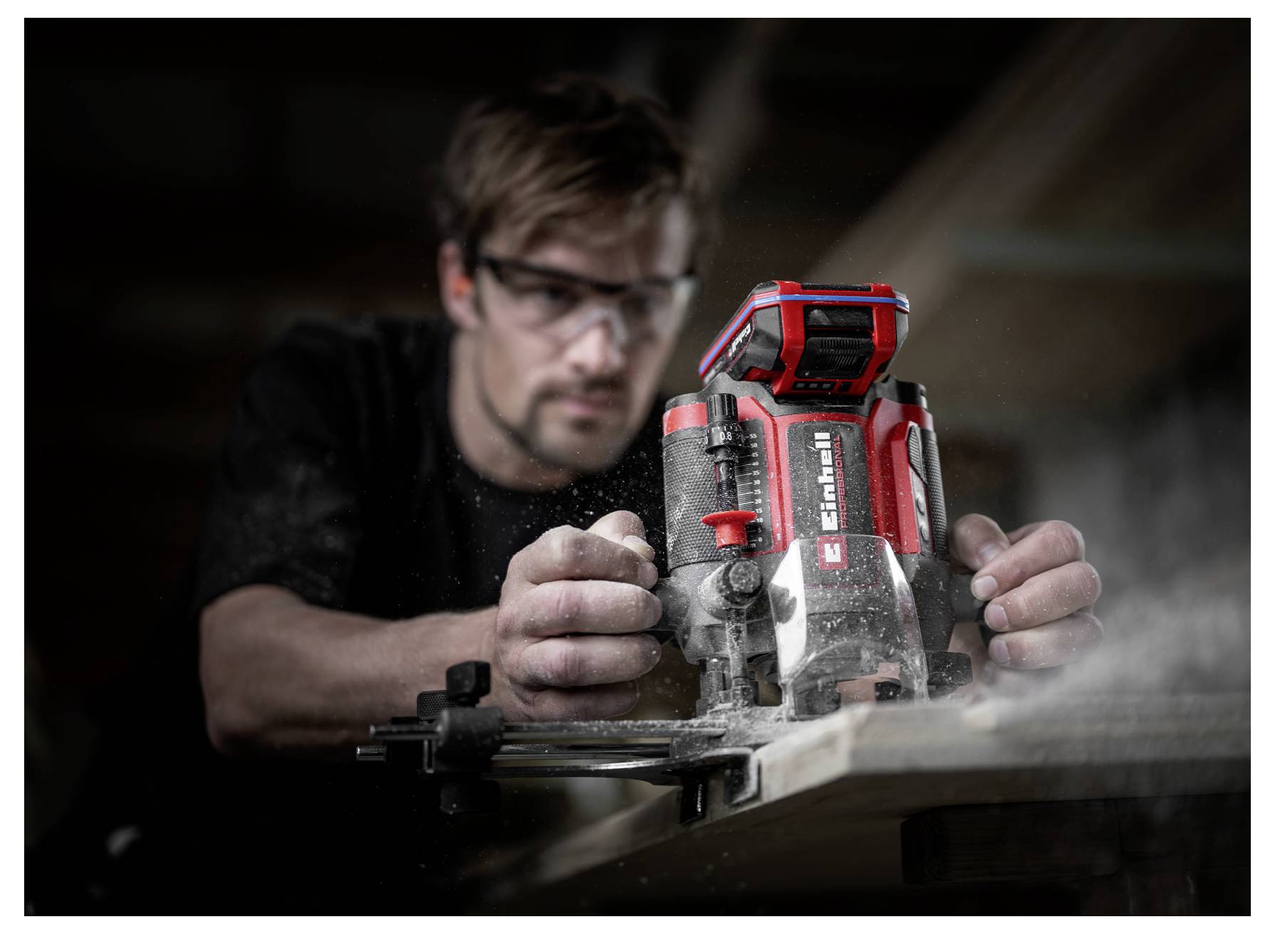 Ein Mann mit einer Schutzbrille verwendet ein rotes tragbares Elektrowerkzeug zum Sägen von Holz, wobei Sägespäne in der Werkstatt in die Luft wirbeln.