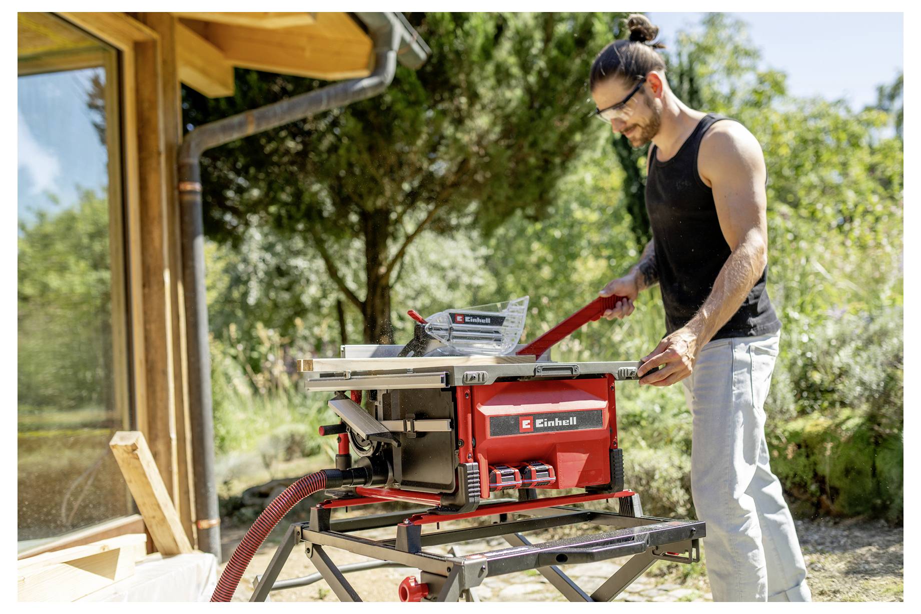 Ein Mann bedient eine rote Tischkreissäge im Freien auf einer Baustelle und schneidet Holz neben einem Haus unter Bäumen.