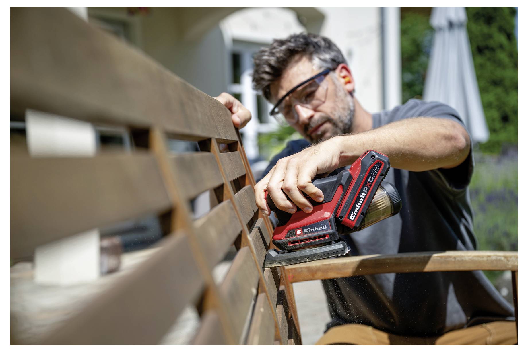 Ein Mann nutzt einen roten Elektroschleiffer, um eine Holzbank im Freien zu glätten. Er trägt eine Schutzbrille und arbeitet an einem sonnigen Tag.