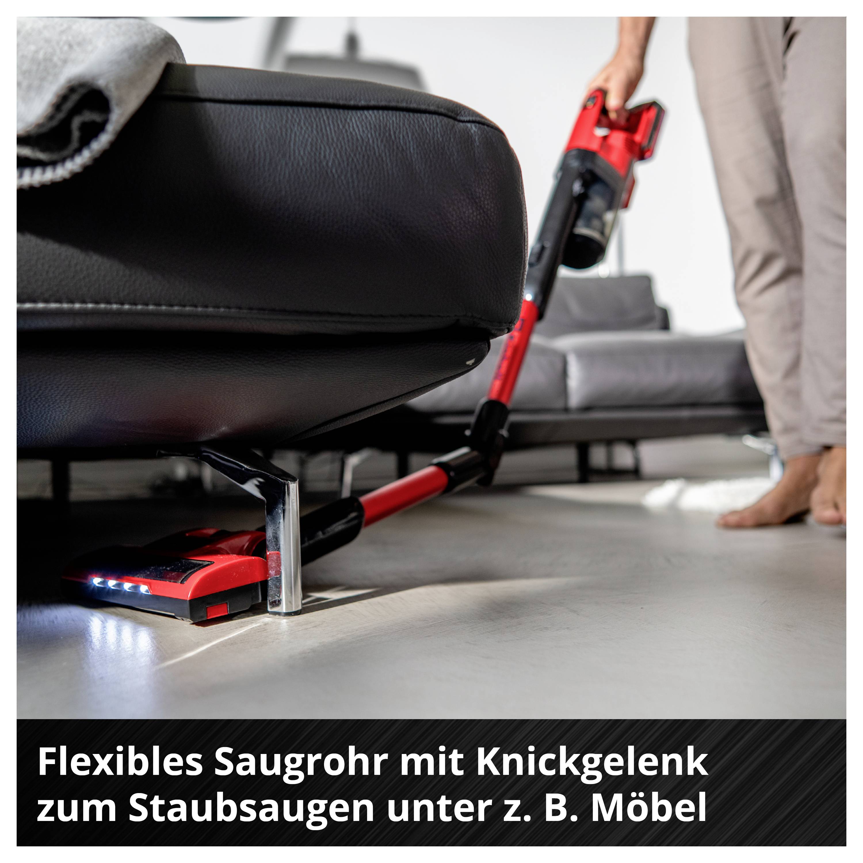 Roter Akkustaubsauger mit Knickgelenk reinigt unter einem Sofa im Wohnzimmer; Hinweis: Flexibles Saugrohr zum Staubsaugen unter Möbeln.