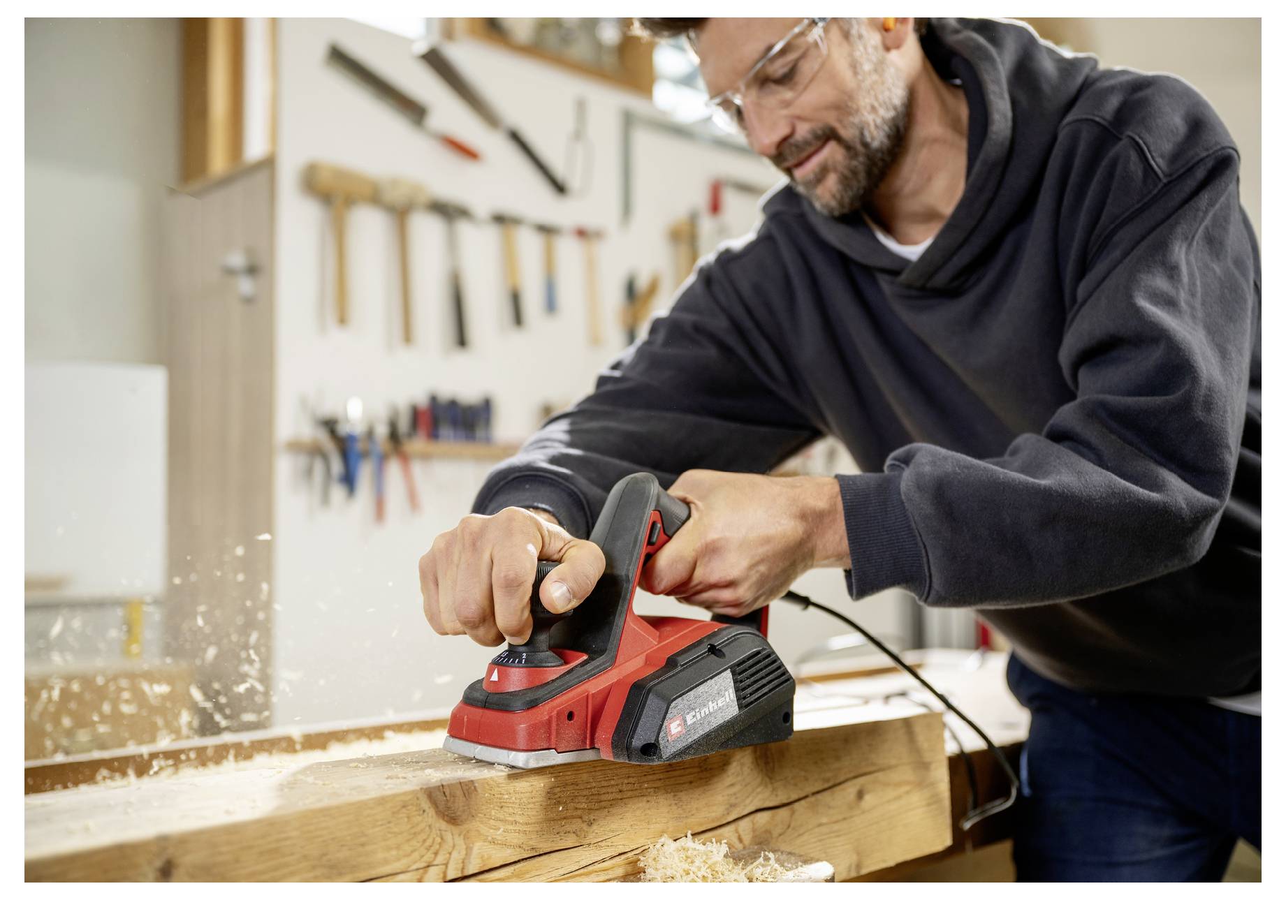 Ein Mann, der einen roten elektrischen Handhobel auf einem Holzbrett in einer Werkstatt benutzt, wobei Holzspäne fliegen.