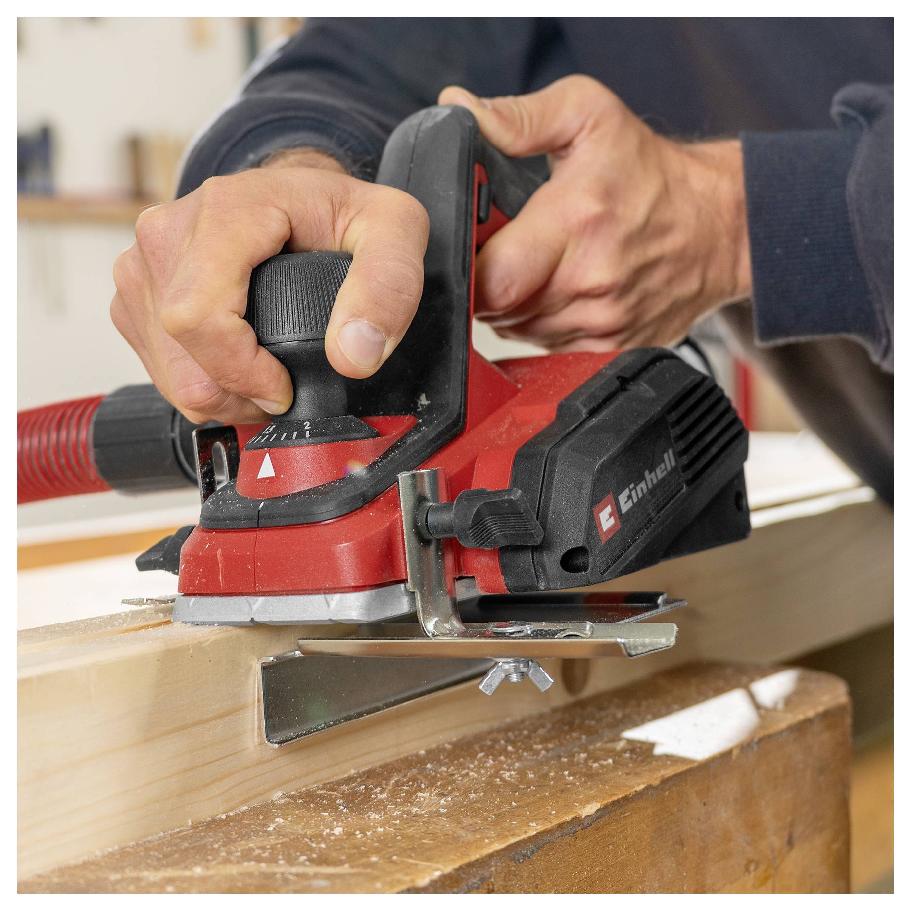Nahaufnahme einer Person, die einen roten elektrischen tragbaren Holzhobelmaschine auf einem Holzbrett verwendet und Holz in einer Werkstatt abspant.