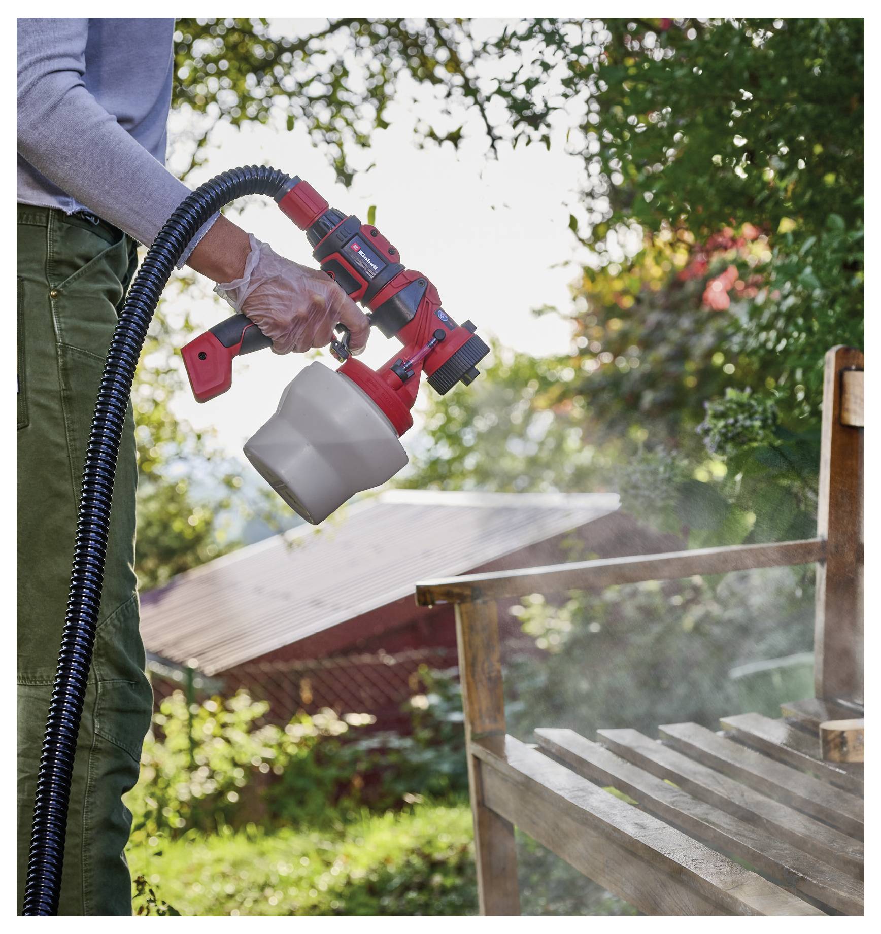 Person mit einem tragbaren Farbsprühgerät, die/der eine hölzerne Outdoor-Bank oder einen Stuhl im Hinterhof mit sichtbarem Sprühnebel beschichtet.