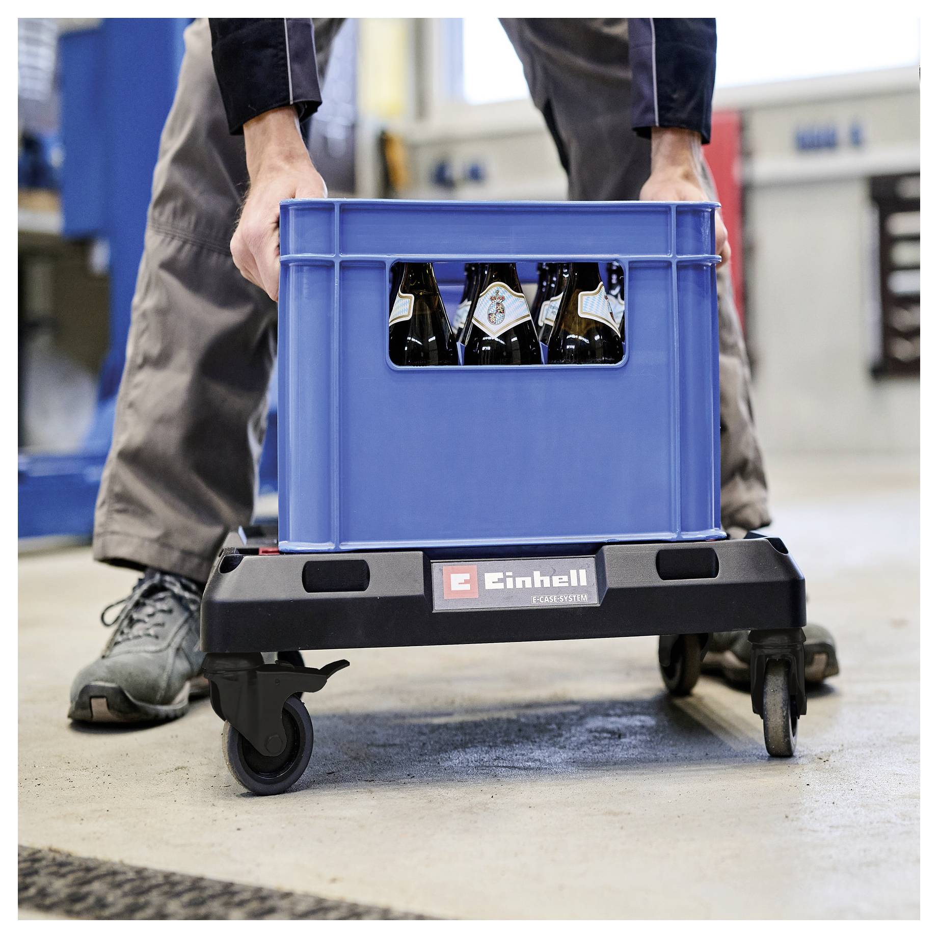 Ein Arbeiter schiebt eine blaue Kiste mit Flaschen auf einem Rad-Einhell-Wagen in einer Industrieumgebung.