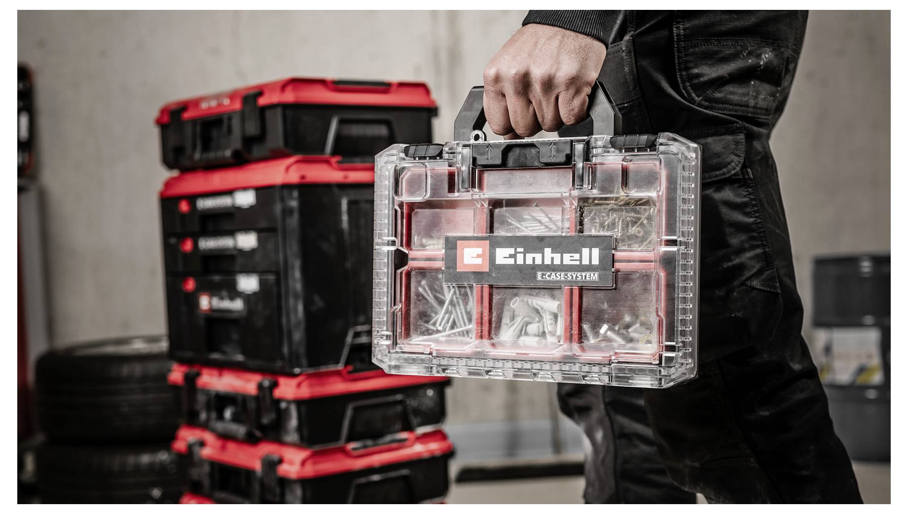 Person in schwarzer Arbeitskleidung hält einen transparenten Kunststoff-Werkzeugorganizer von Einhell mit kleinen Teilen, neben gestapelten rot-schwarzen Lagerkästen.