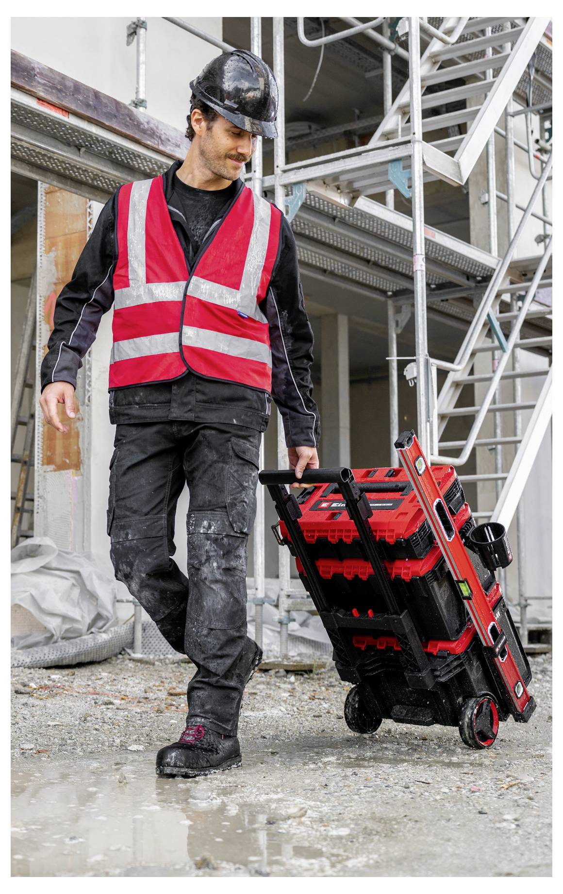 Bauarbeiter in Schutzhelm und Warnweste zieht eine Werkzeugkiste mit Rädern auf einer Baustelle.