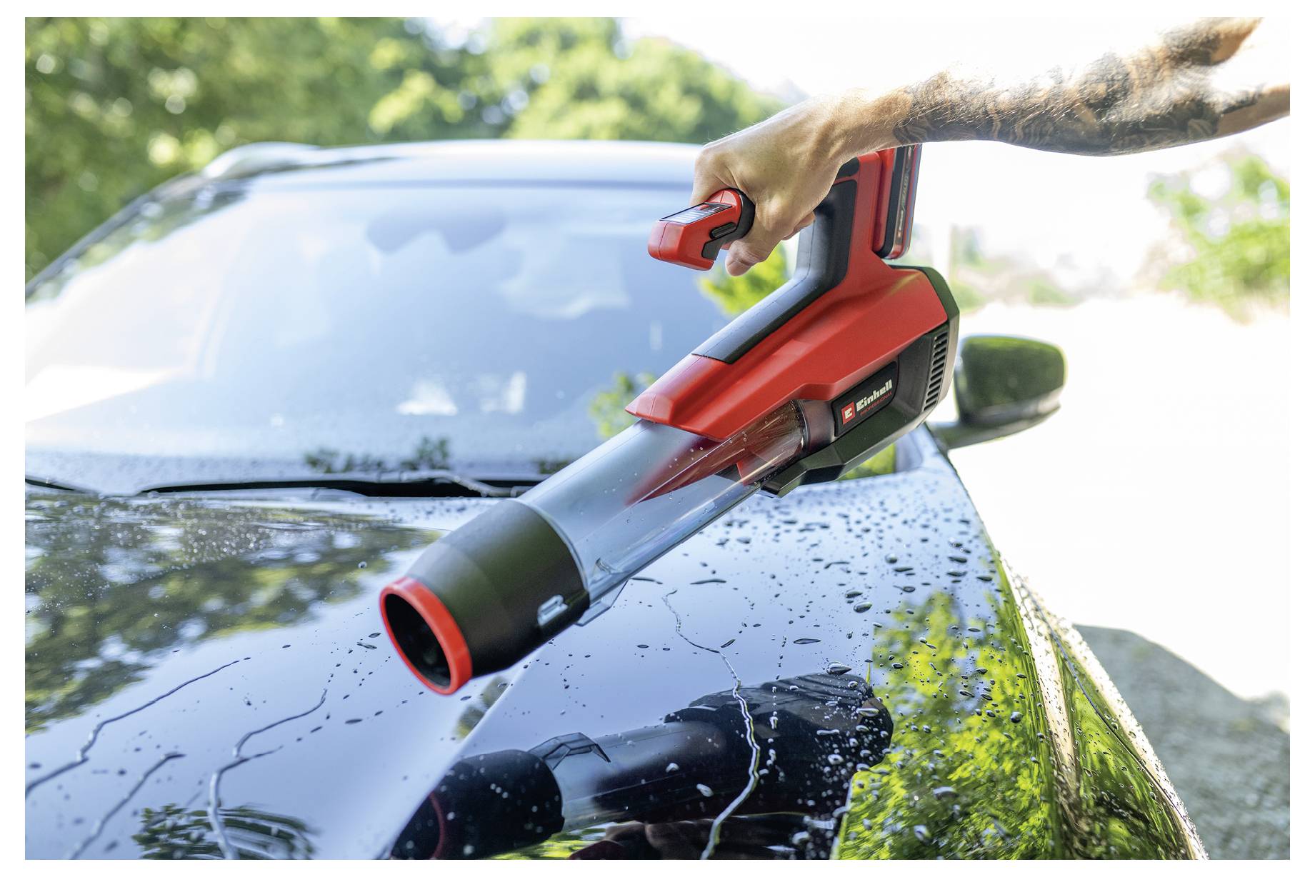 Eine Person, die ein rotes schnurloses Laubgebläse verwendet, um Wasser und Schmutz von einer nassen Motorhaube eines Autos im Freien zu entfernen.