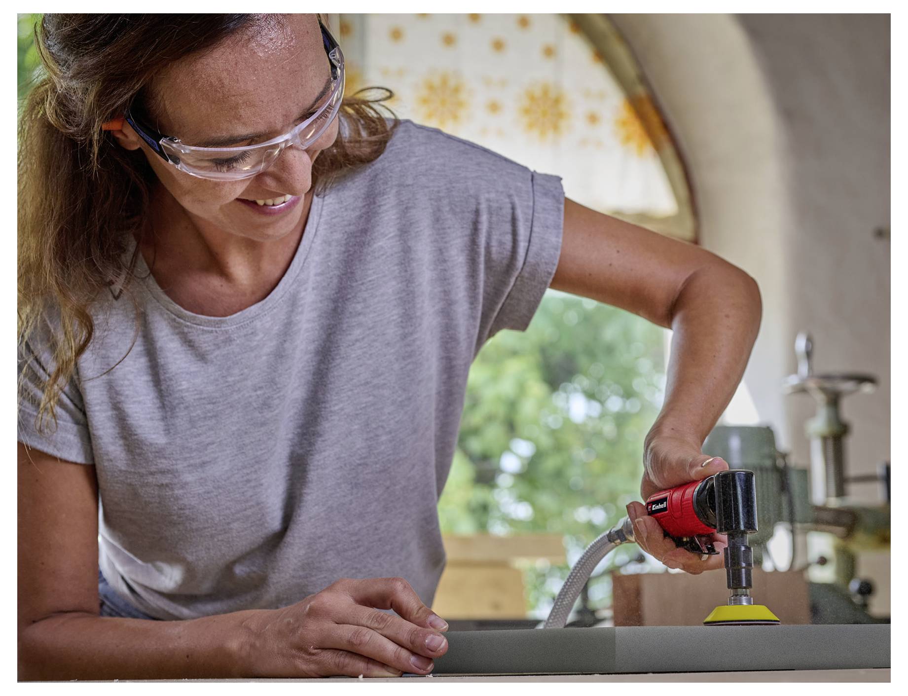 Eine Frau, die Schutzbrille trägt, schleift eine Oberfläche mit einem Elektrowerkzeug in einer Werkstatt.