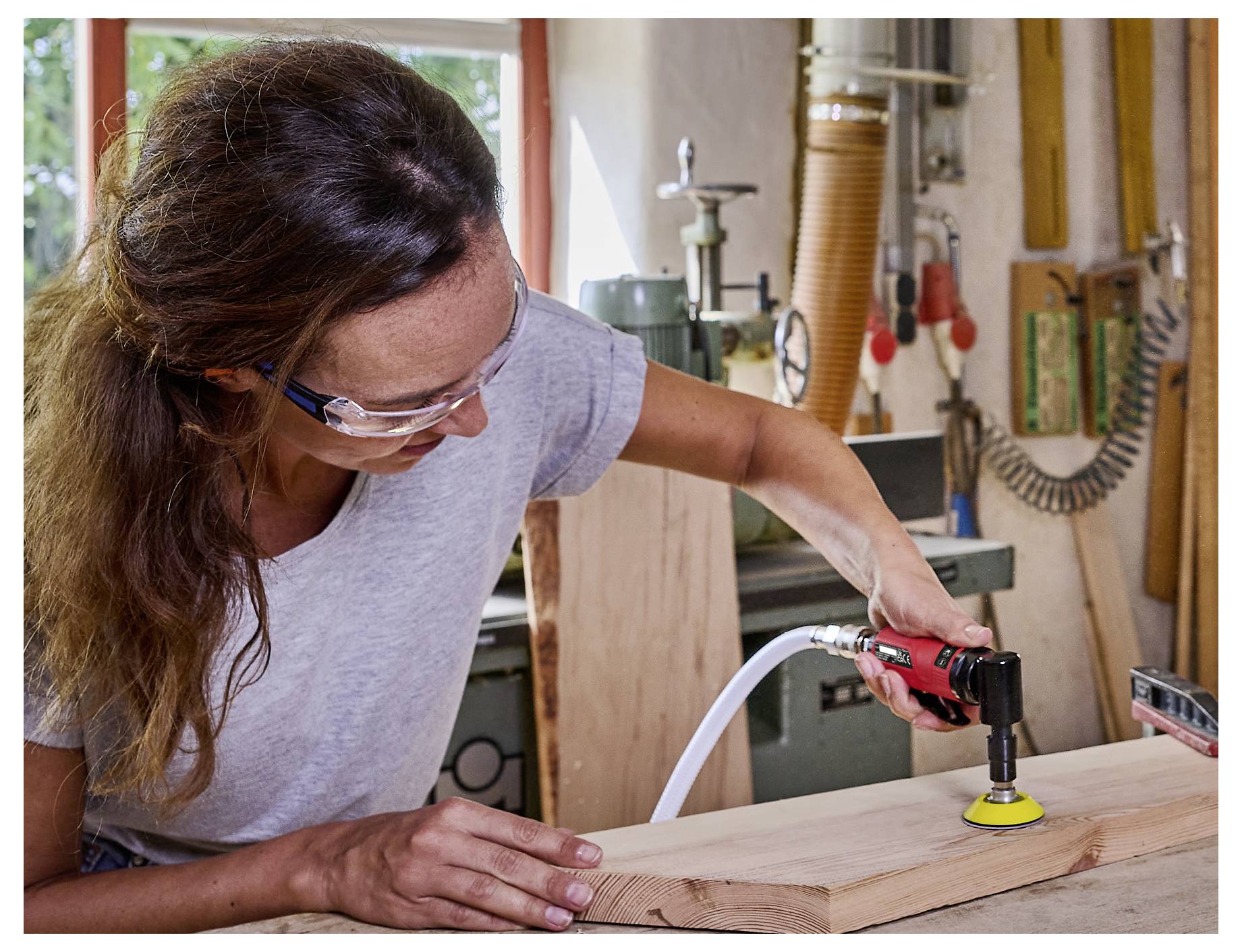 Eine Person verwendet einen Elektroschleiffer auf einer Holzplatte in einer Werkstatt, trägt eine Schutzbrille und ein graues Hemd.