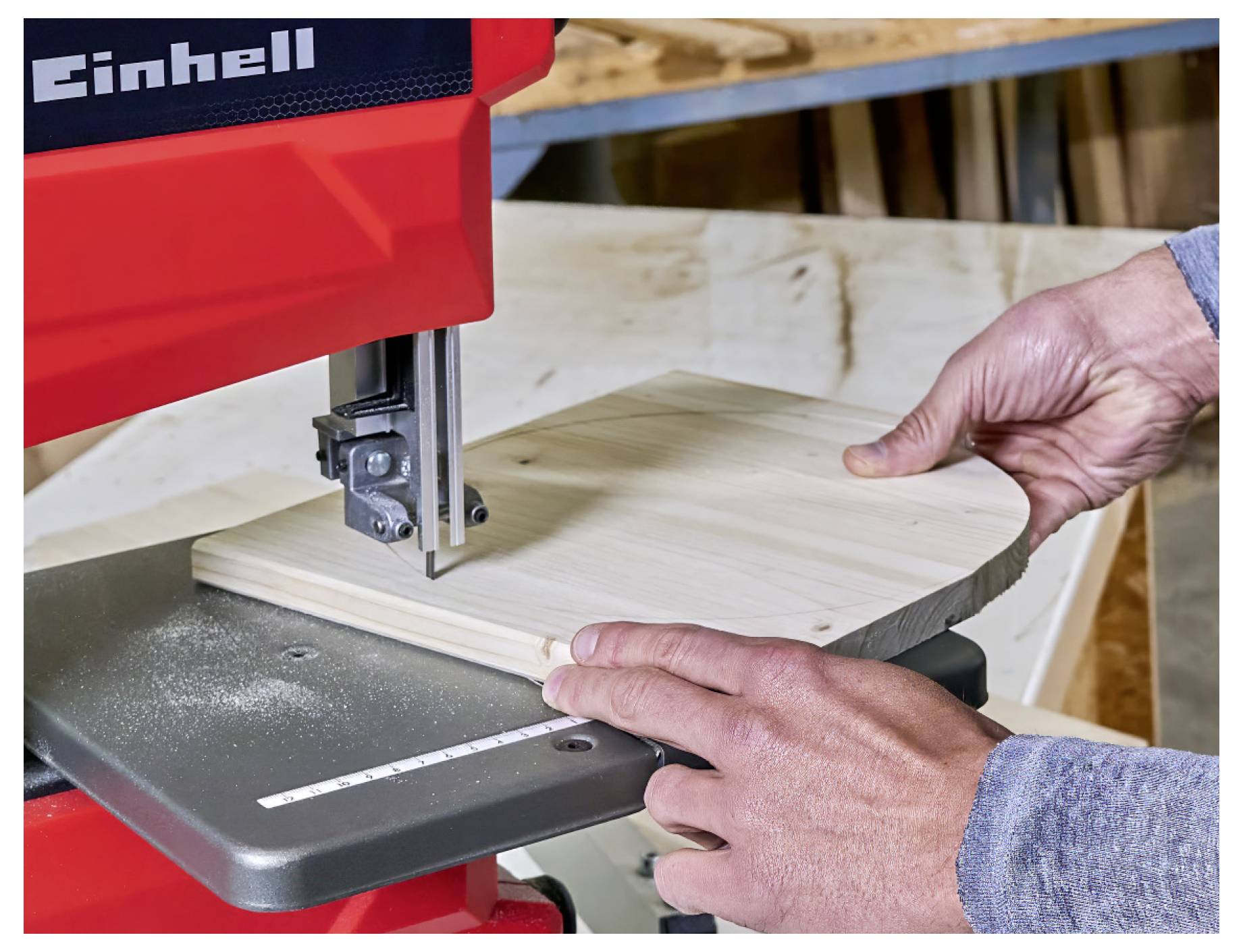 Hände führen ein Holzbrett durch eine rote Einhell-Bandsaw in einer Werkstatt und schneiden eine gekrümmte Form.