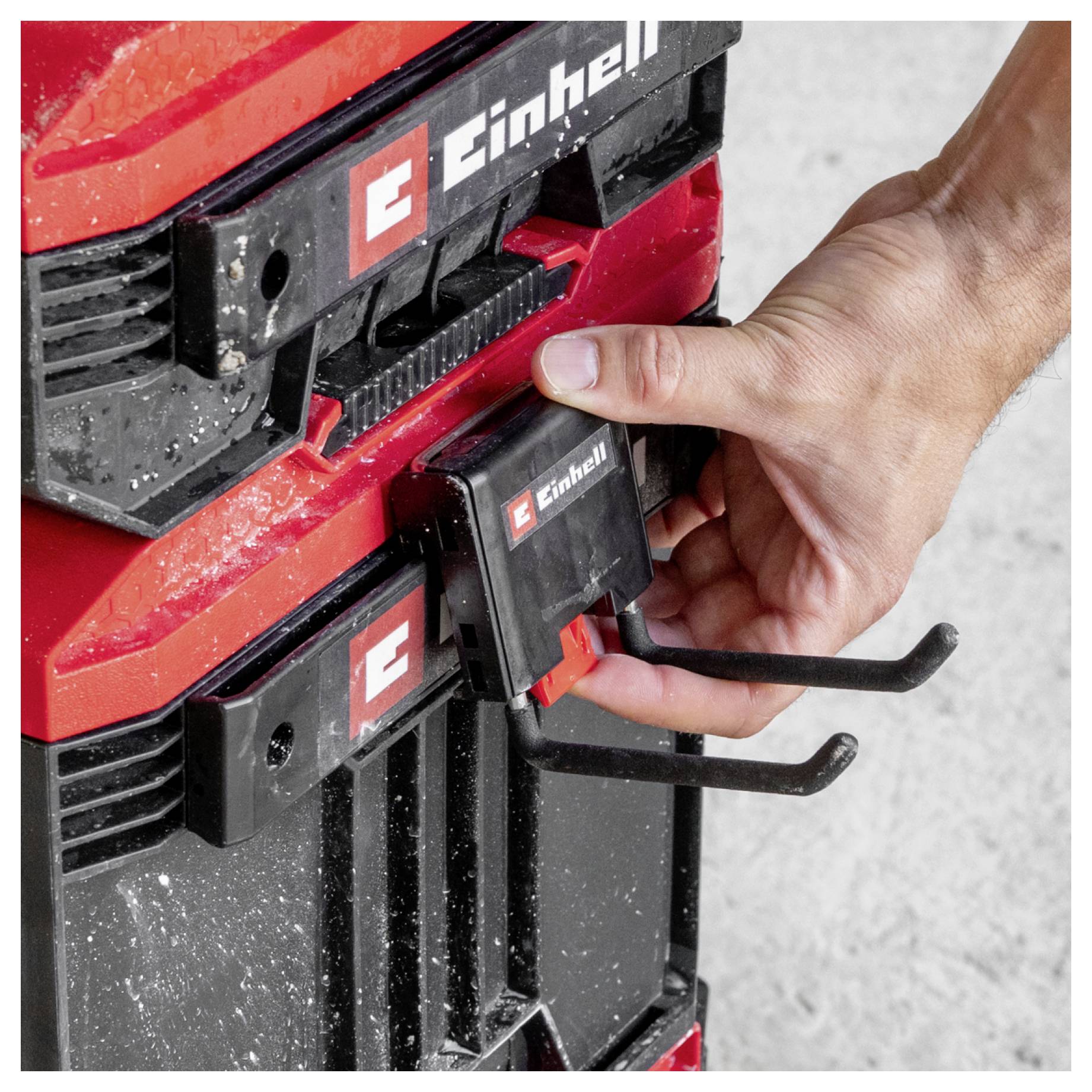 Hand entnimmt einen Einhell-Akku aus einem roten Elektrowerkzeug oder Akkuladegerät, wobei zwei schwarze Metallhaken sichtbar sind.