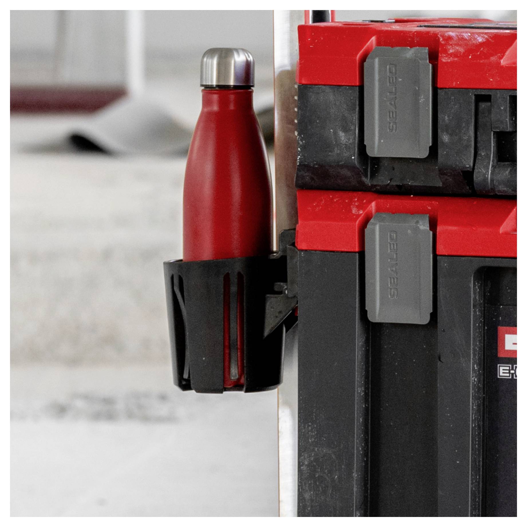Rote Wasserflasche in einem schwarzen Halter befestigt, der an gestapelten rot-schwarzen Werkzeugkästen oder Lagerkästen angebracht ist.