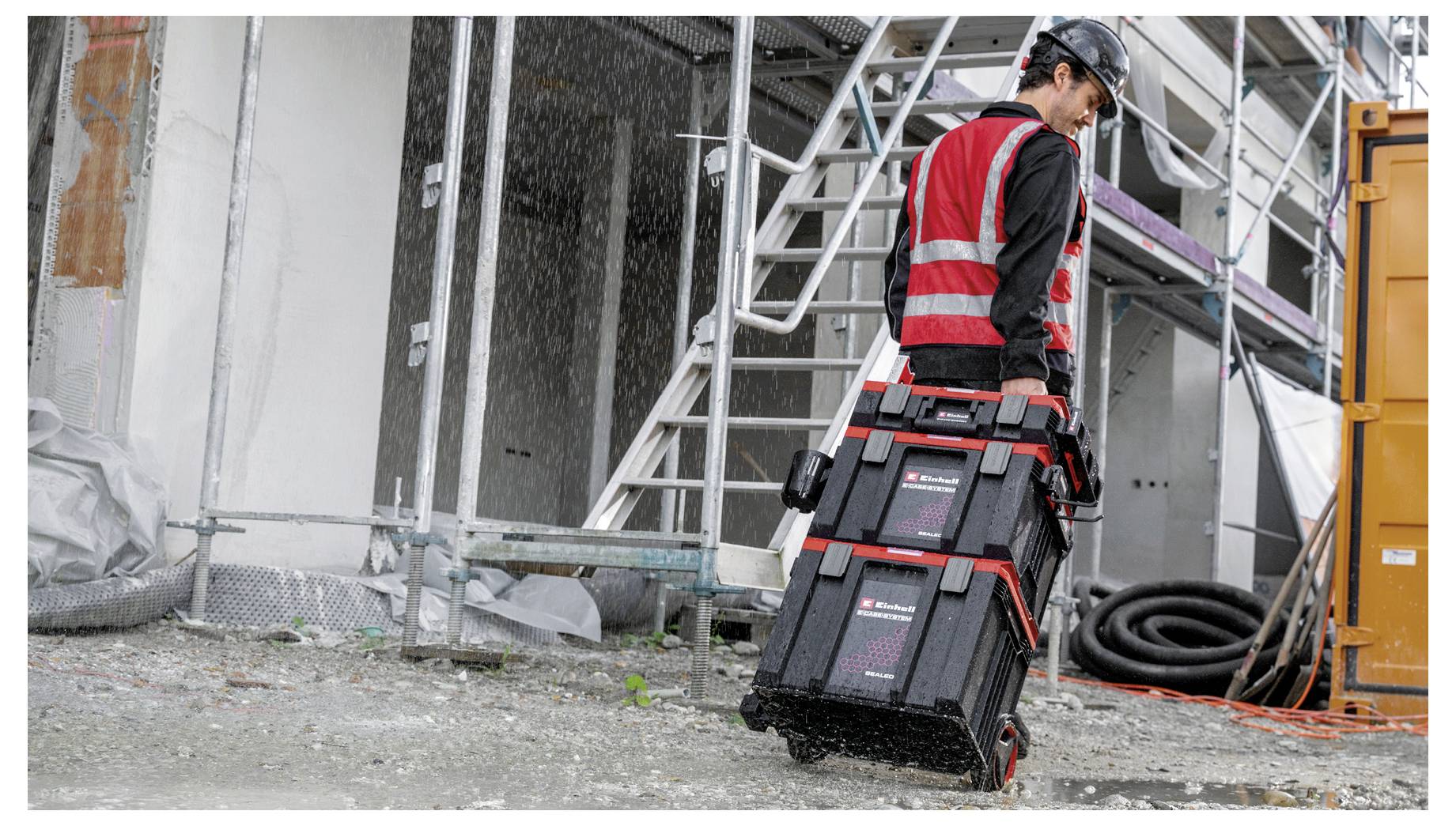 Ein Bauarbeiter in einer roten Sicherheitsweste und einem Schutzhelm trägt gestapelte Werkzeugkisten über eine schlammige Baustelle neben Gerüsten und einem orangefarbenen Container.