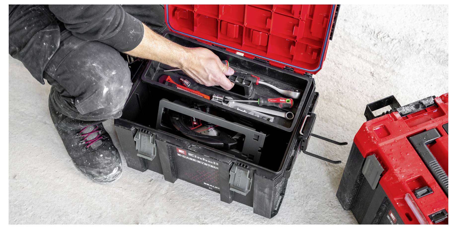 Person kneeling by an open black tool case, reaching for tools inside, with a red toolbox beside it on a concrete floor.