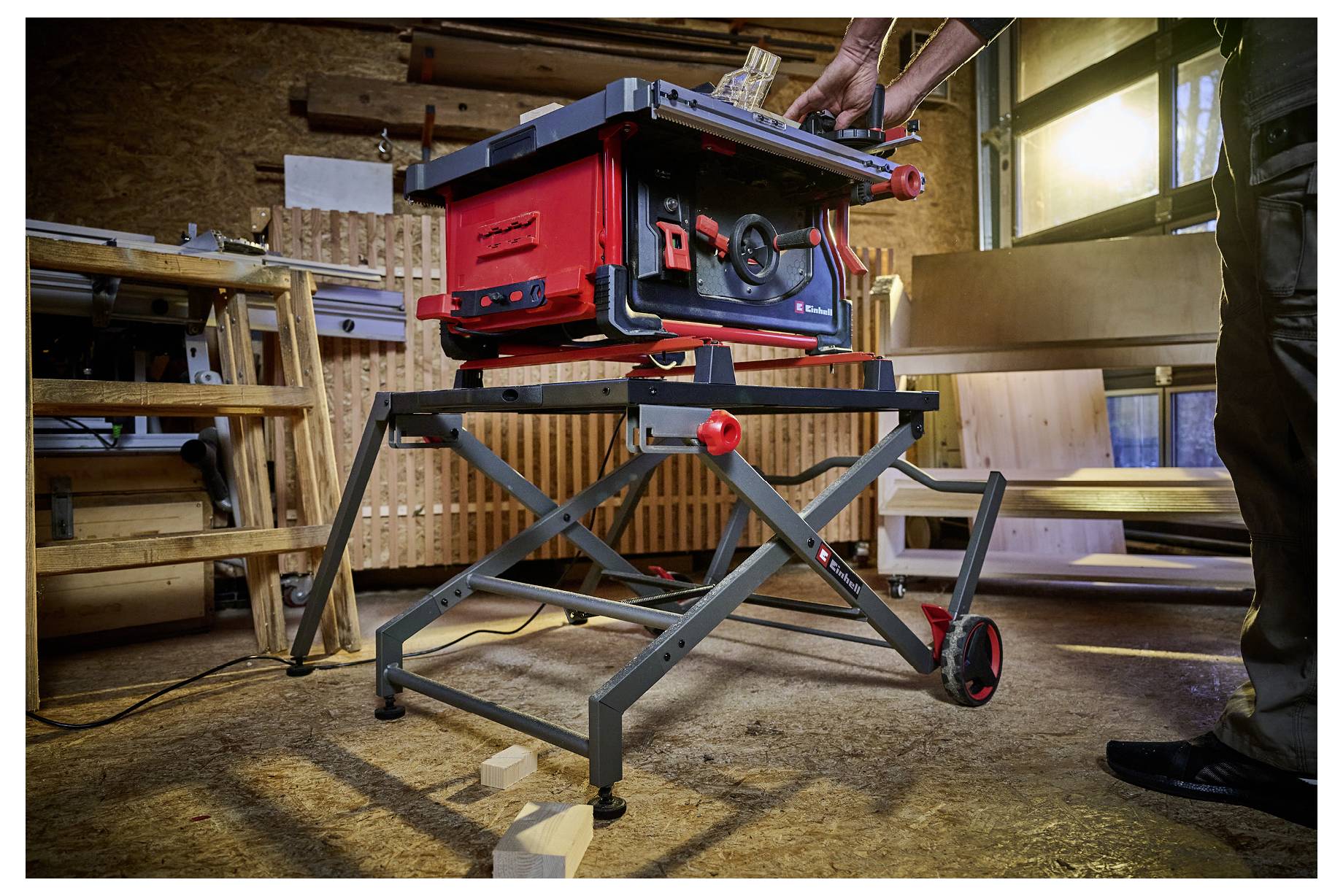 Eine Person stellt eine rote Tischkreissäge auf einem Klapptisch in einer Werkstatt ein, mit Holzbrettern, einer Leiter und Sonnenlicht durch ein Fenster.