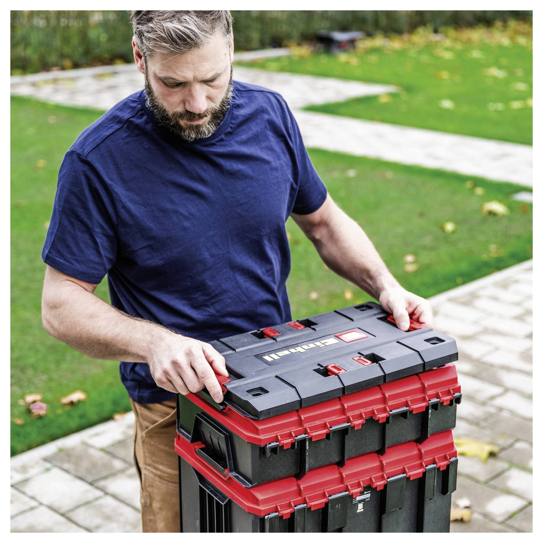 A man outdoors opens a stacked black-and-red plastic tool box on a stone patio, inspecting it in a yard.