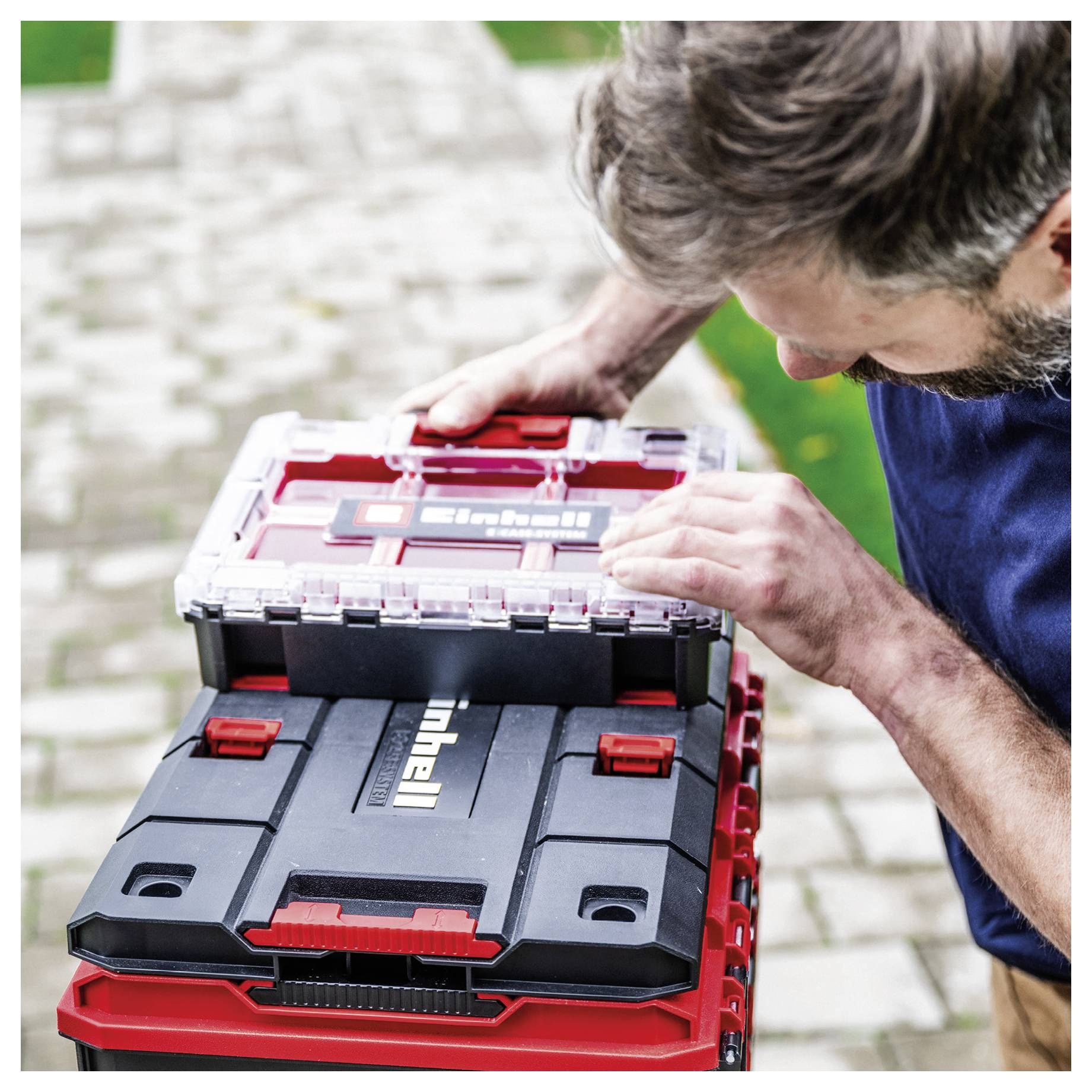 Man examining a stack of red and black toolboxes, checking the top case outdoors on a paved path.