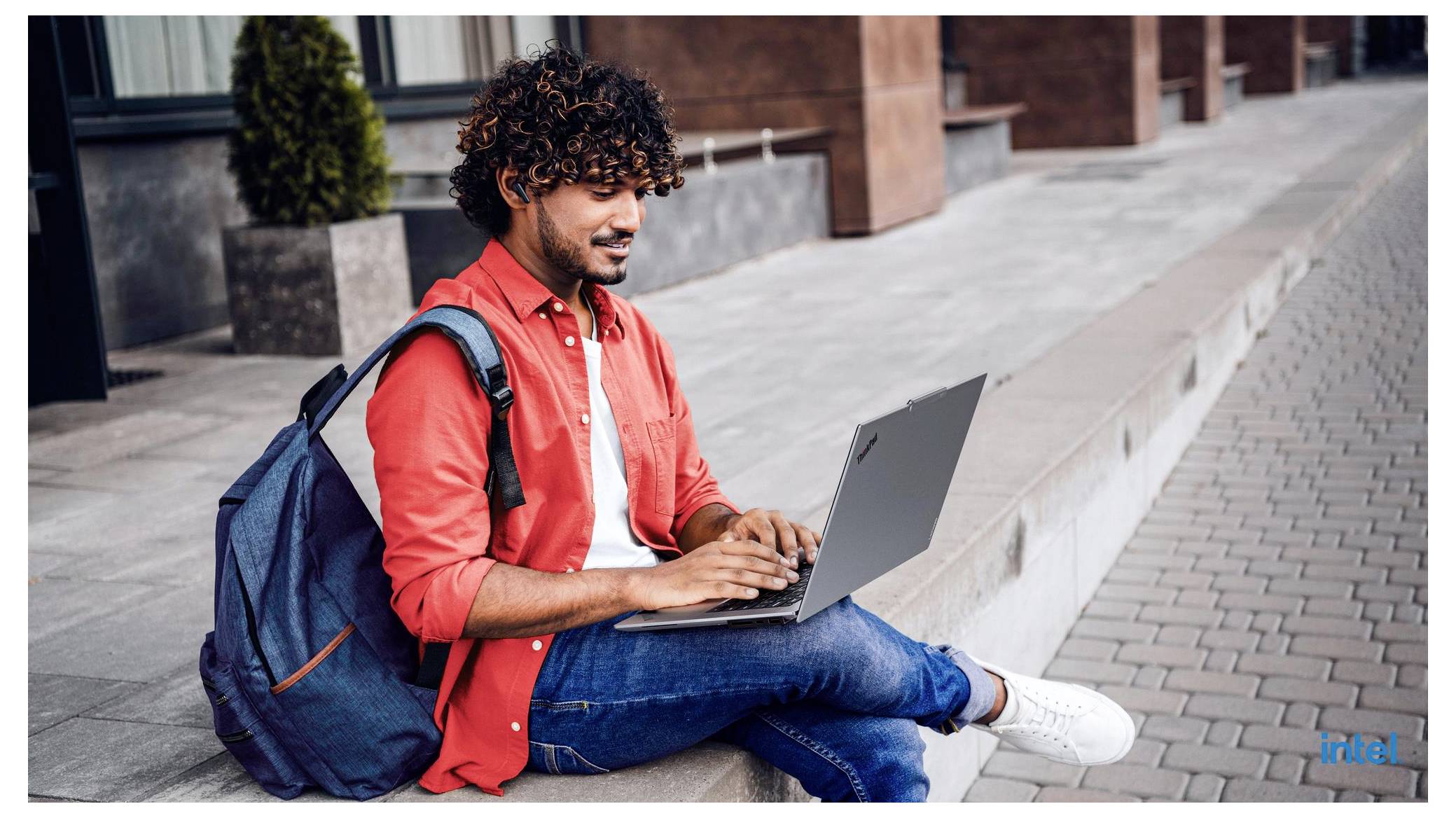 Ein Mann in einem roten Hemd sitzt draußen mit einem Rucksack und nutzt einen Laptop auf dem Gehweg neben einem Gebäude.