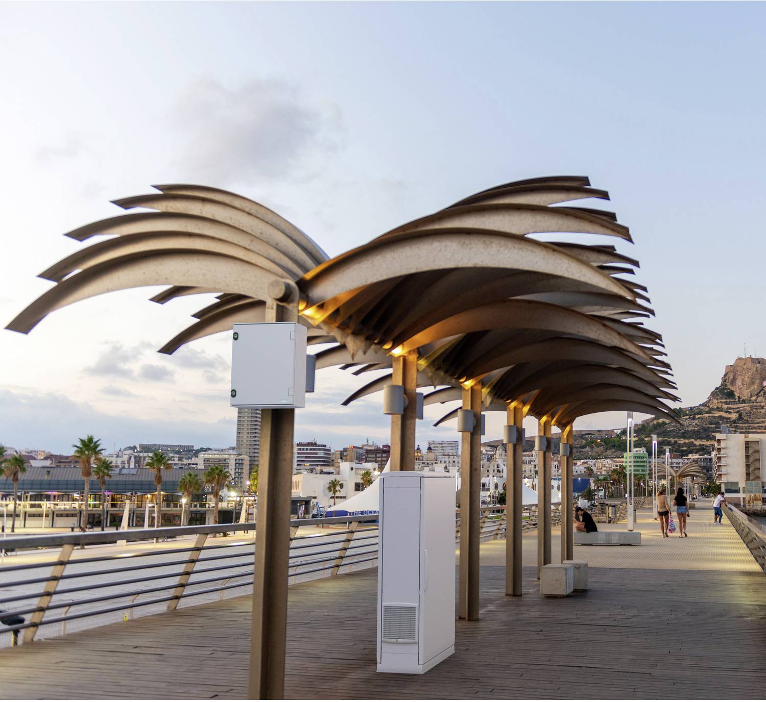 Moderne Strandpromenade mit einer Reihe von beleuchteten, palmenförmigen Überdachungen entlang einer Uferpromerade bei Dämmerung, mit Stadtgebäuden und einem Hügel im Hintergrund.
