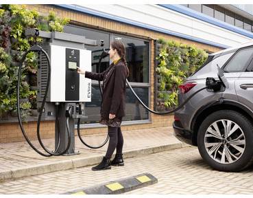 Frau lädt ein Elektroauto an einer öffentlichen Ladenstation außerhalb eines Gebäudes auf, wobei das Kabel mit dem Fahrzeug verbunden ist.