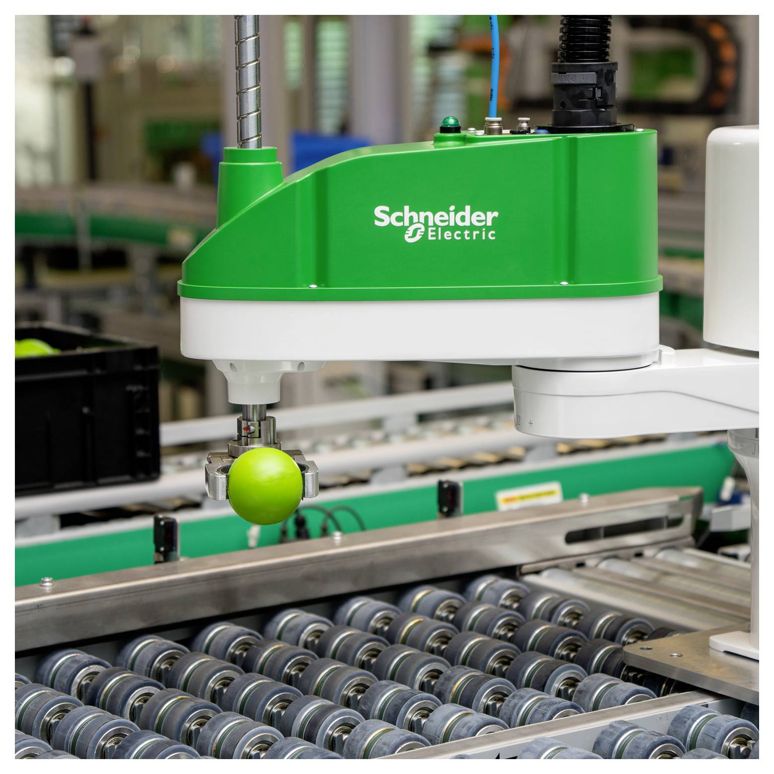 Schneider Electric Roboterarm nimmt einen grünen Apfel über einem Fabrikförderband mit Metallrollen auf.