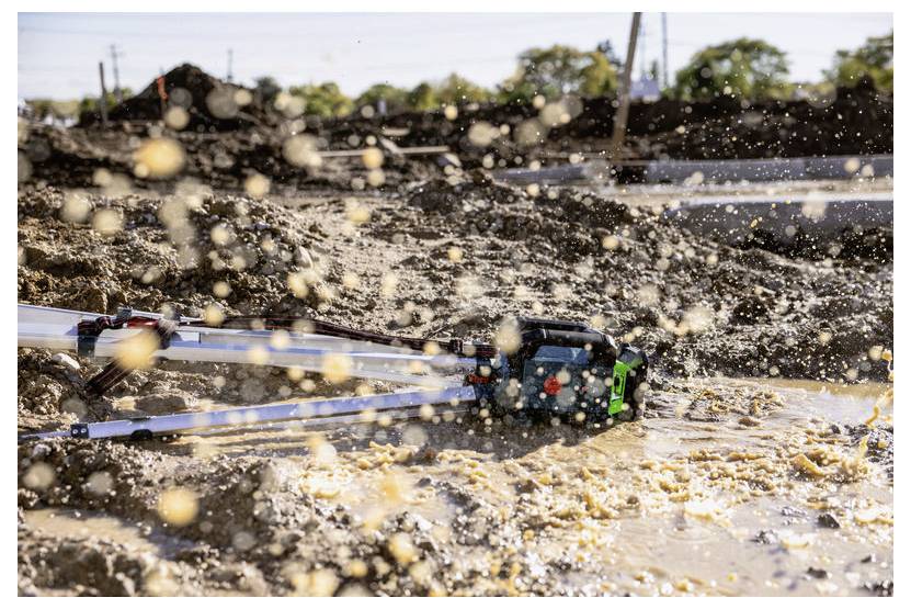 Baggerschaufel rührt schlammiges Wasser auf einer Baustelle auf, wobei Schmutz über den Vordergrund spritzt.