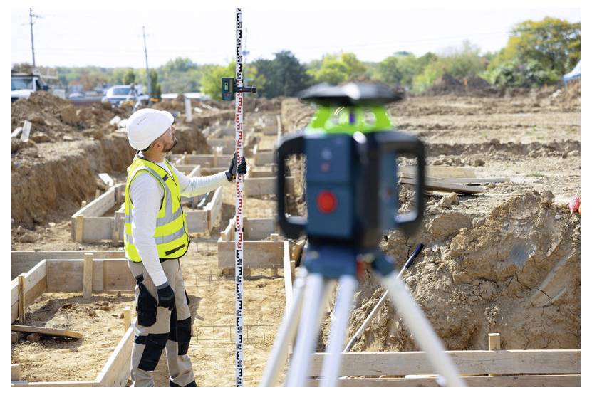 Vermesser in Schutzhelm und Sicherheitsweste misst eine Baustelle mit einem Messstab, während ein auf einem Stativ montiertes Laser-Nivelliergerät im Vordergrund zu sehen ist.