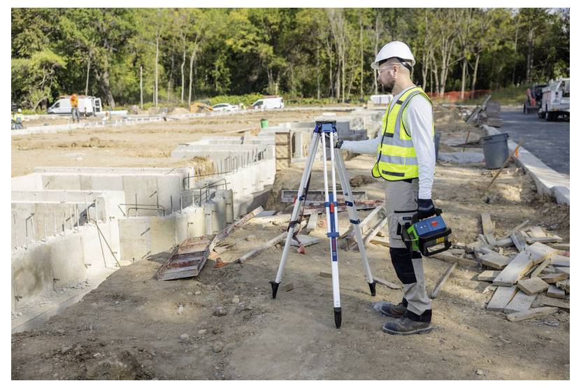 Vermesser in Schutzhelm und Sicherheitsweste, der ein Dreibein-montiertes Messinstrument auf einer Straßenbaustelle verwendet.