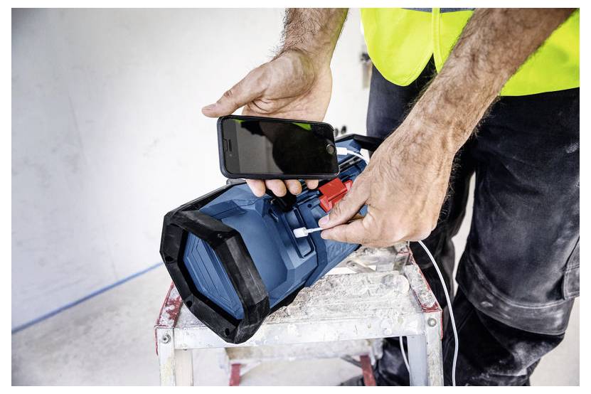 Person in einem leuchtenden Warnweste verwendet ein Smartphone, um einen blauen tragbaren Lautsprecher auf einer Werkbank aufzuladen oder zu verbinden.