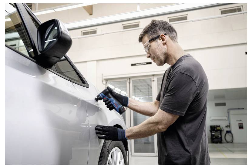 Mechaniker in Handschuhen inspiziert eine silberne Autotür mit einem tragbaren Gerät in einer Autowerkstatt.