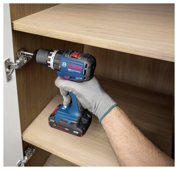 Behandschuhte Hand, die einen Akku-Bohrer verwendet, um ein Schrankenscharnier in einem Holzschrank zu installieren oder festzuziehen.