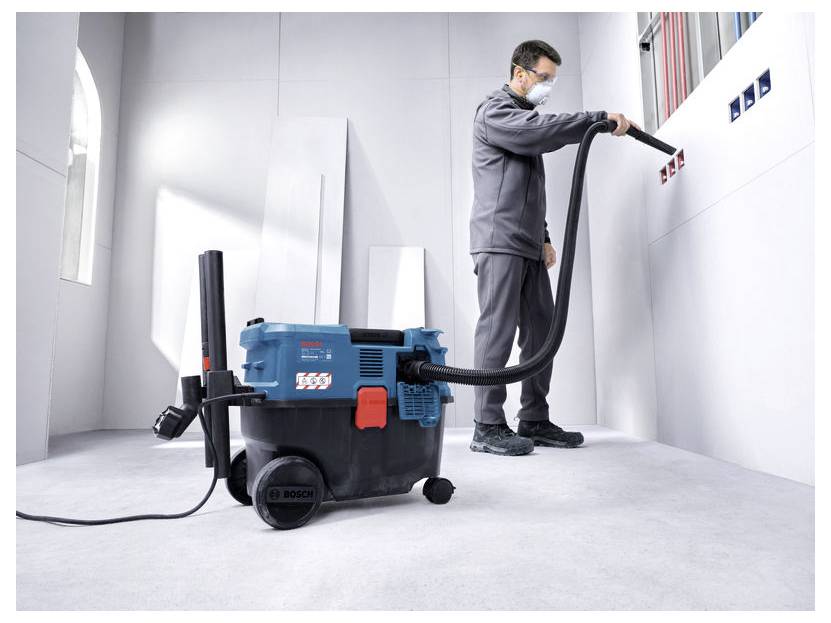 Ein Mann mit Maske benutzt einen großen Bosch Nass-/Trockensauger, um Staub von einer Wand in einem leeren Raum, der sich in Renovierung befindet, zu reinigen.