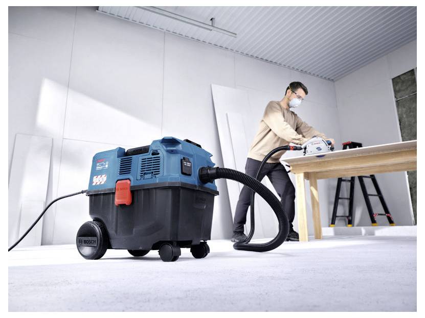 Ein Mann benutzt ein Elektrowerkzeug an einer Werkbank, während ein Bosch Nass-Trockensauger mit Schlauch im Vordergrund auf einer Baustelle steht.