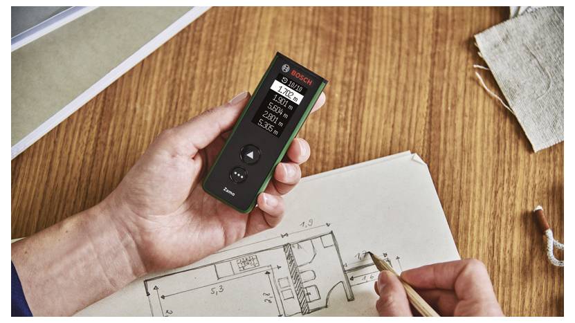Hand, die einen Bosch-Laser-Entfernungsmesser über architektonische Grundrisse hält, während eine andere Hand Messwerte auf Papier skizziert.