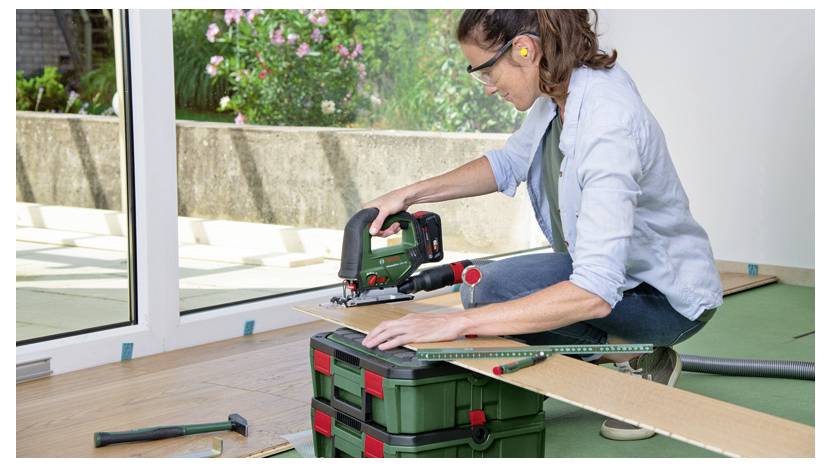 Eine Person, die eine Stichsäge verwendet, um ein Holzbrett auf einer Werkbank im Freien in einem Garten- oder Terrassenbereich zu schneiden.